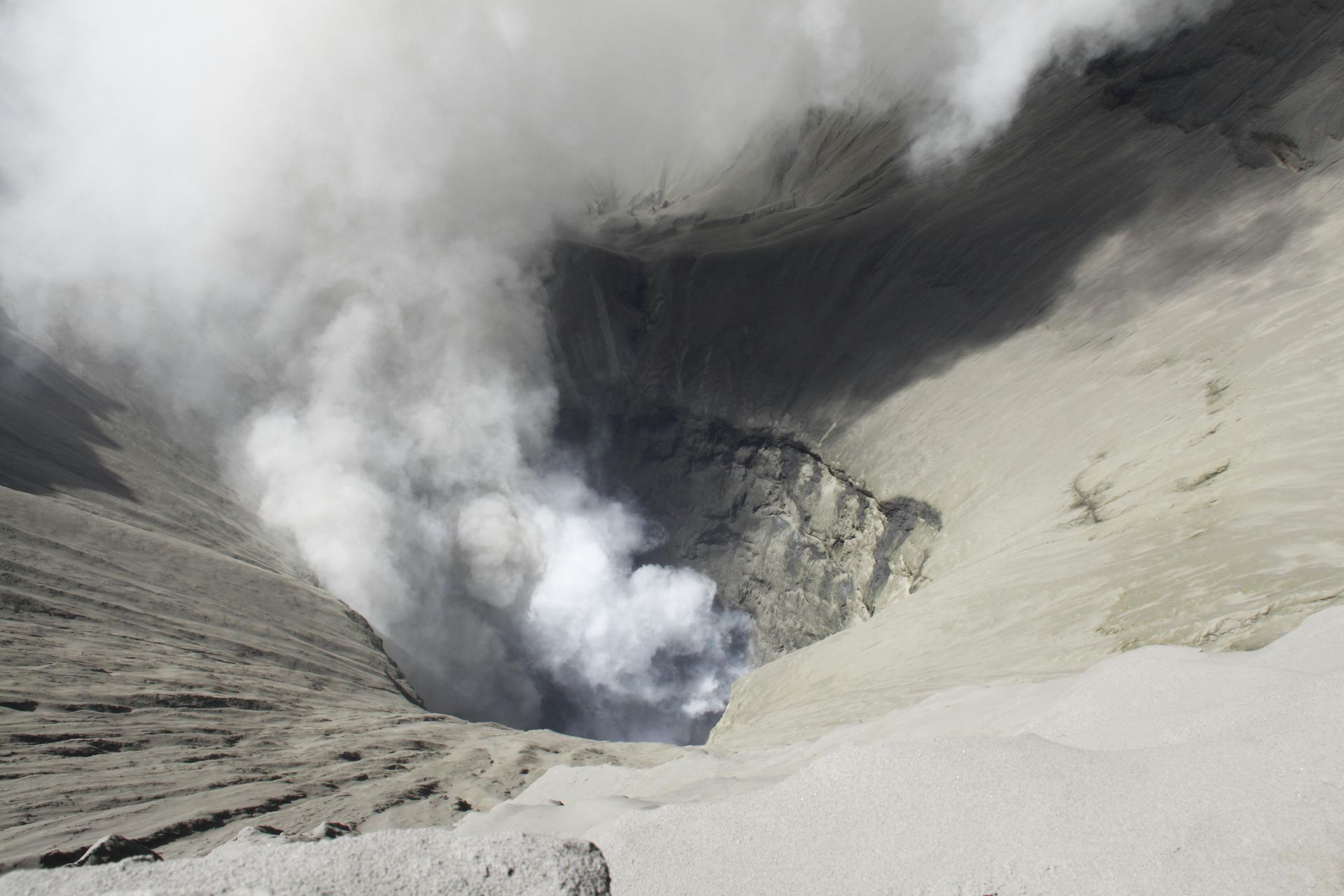 Volcanic crater emitting white smoke; gray ash surrounds it.
