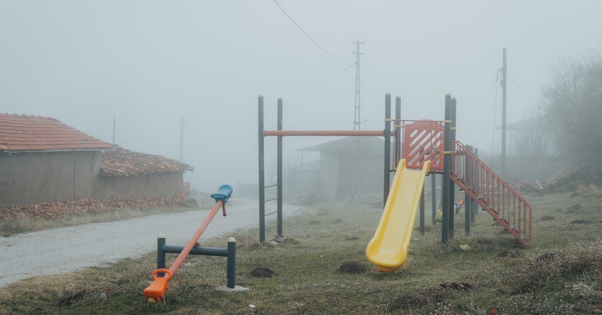 Playground in heavy fog: slide, seesaw, and climbing bars stand empty next to a small building.
