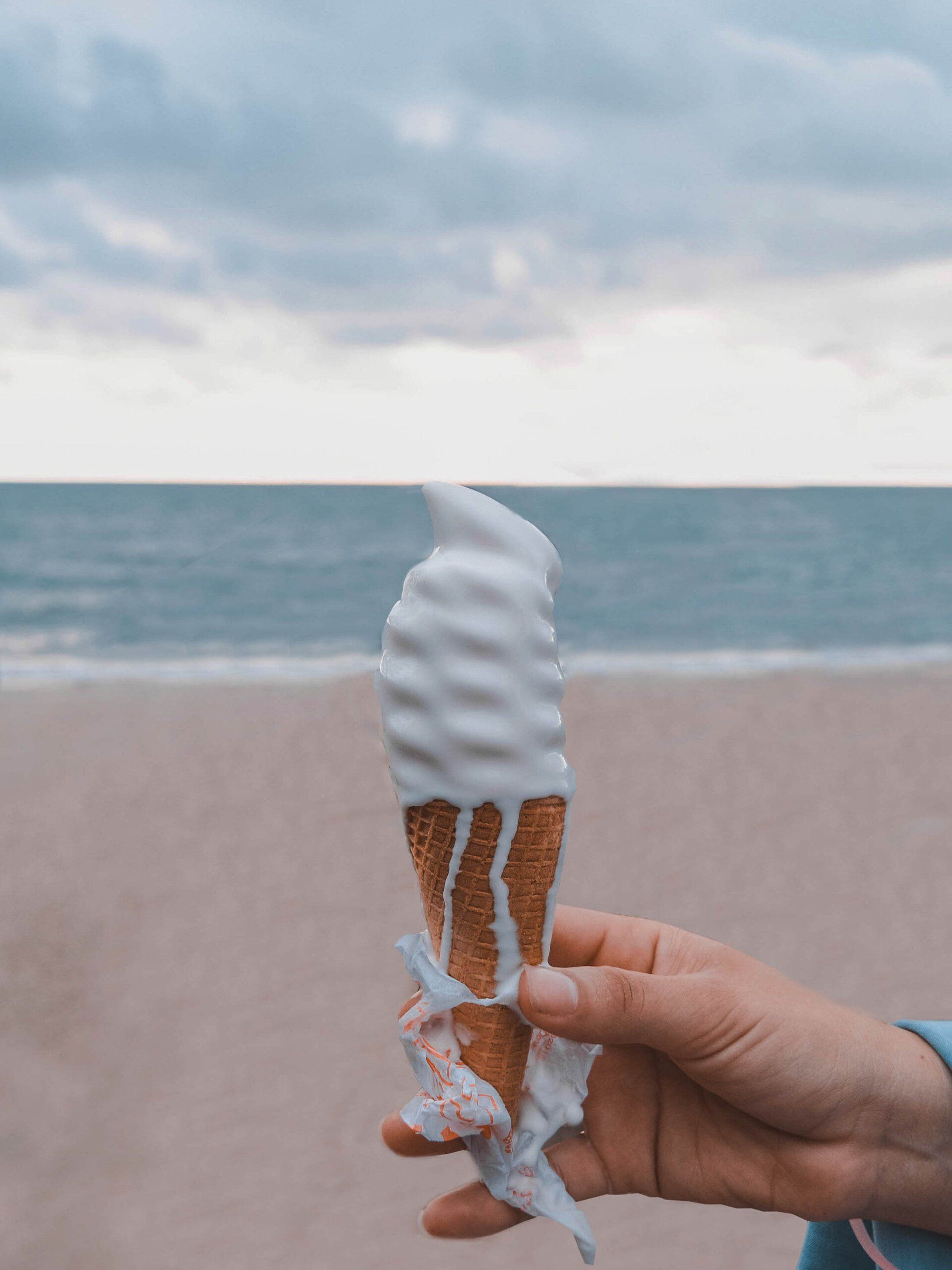 Hand holding a melting ice cream cone on a beach, ocean in background.