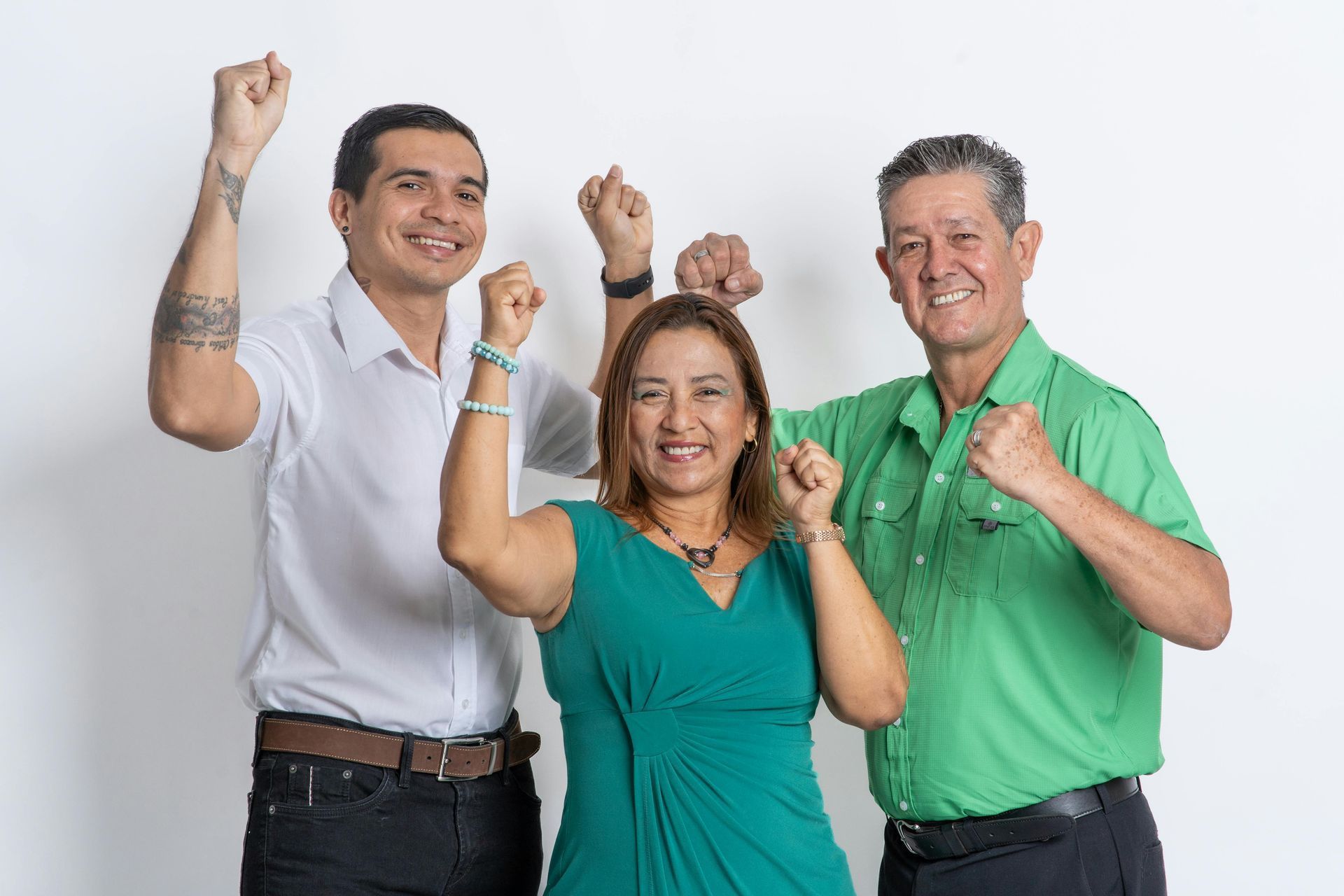 Three people celebrating, arms raised. Man with tattoos smiles; woman in teal dress; man in green shirt. White background.