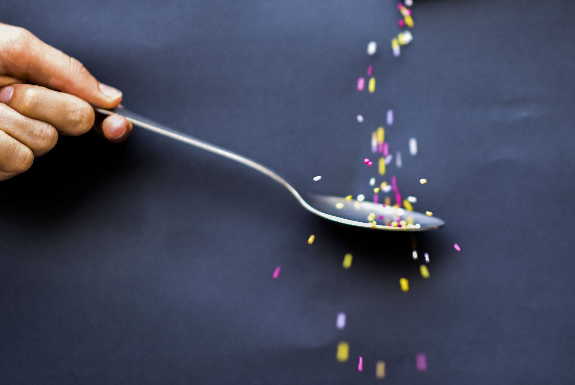 Hand holding a spoon, pouring colorful sprinkles onto it against a dark background.