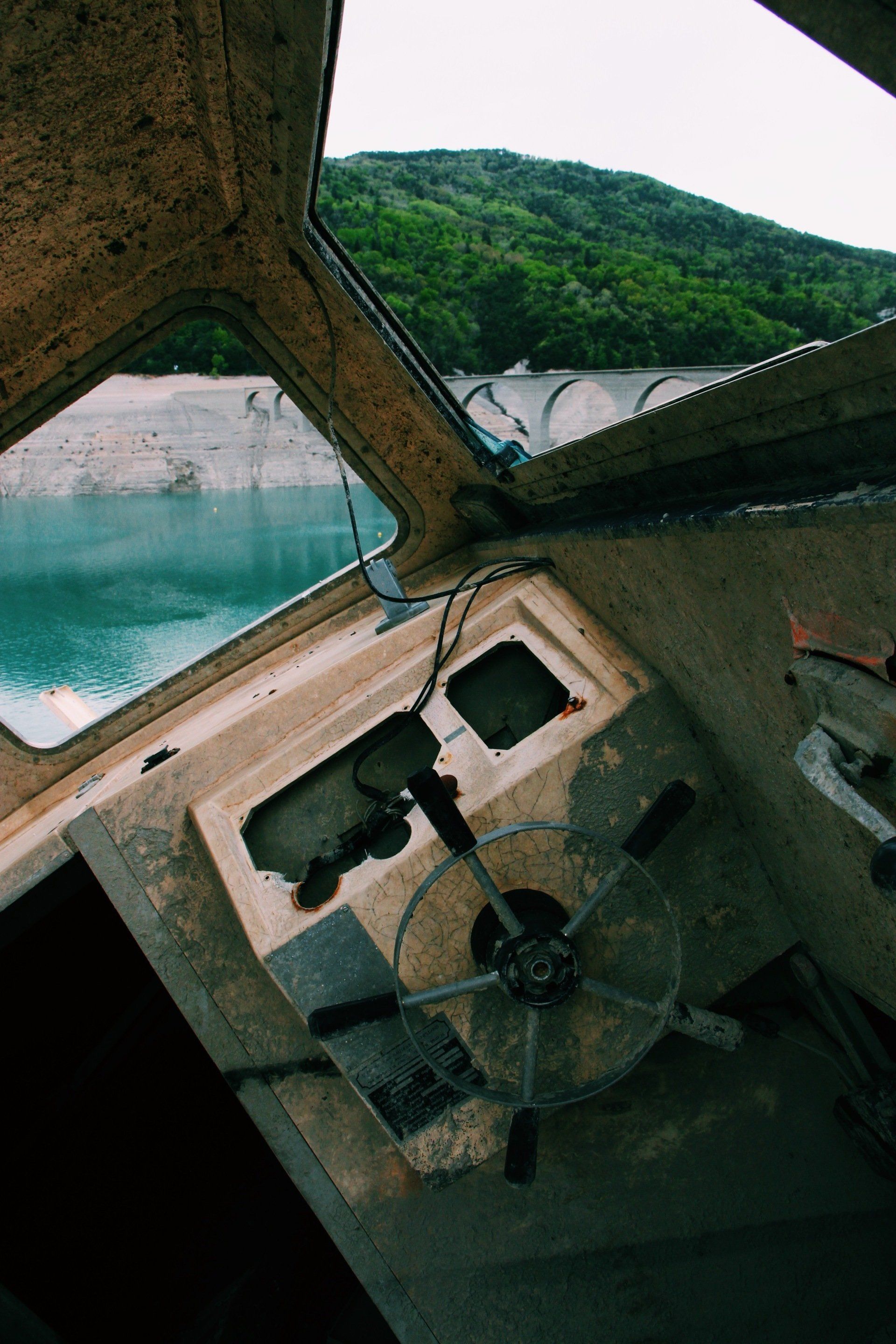 Interior view of an abandoned boat overlooking a lake and dam green hillside & tourquoise water visible 