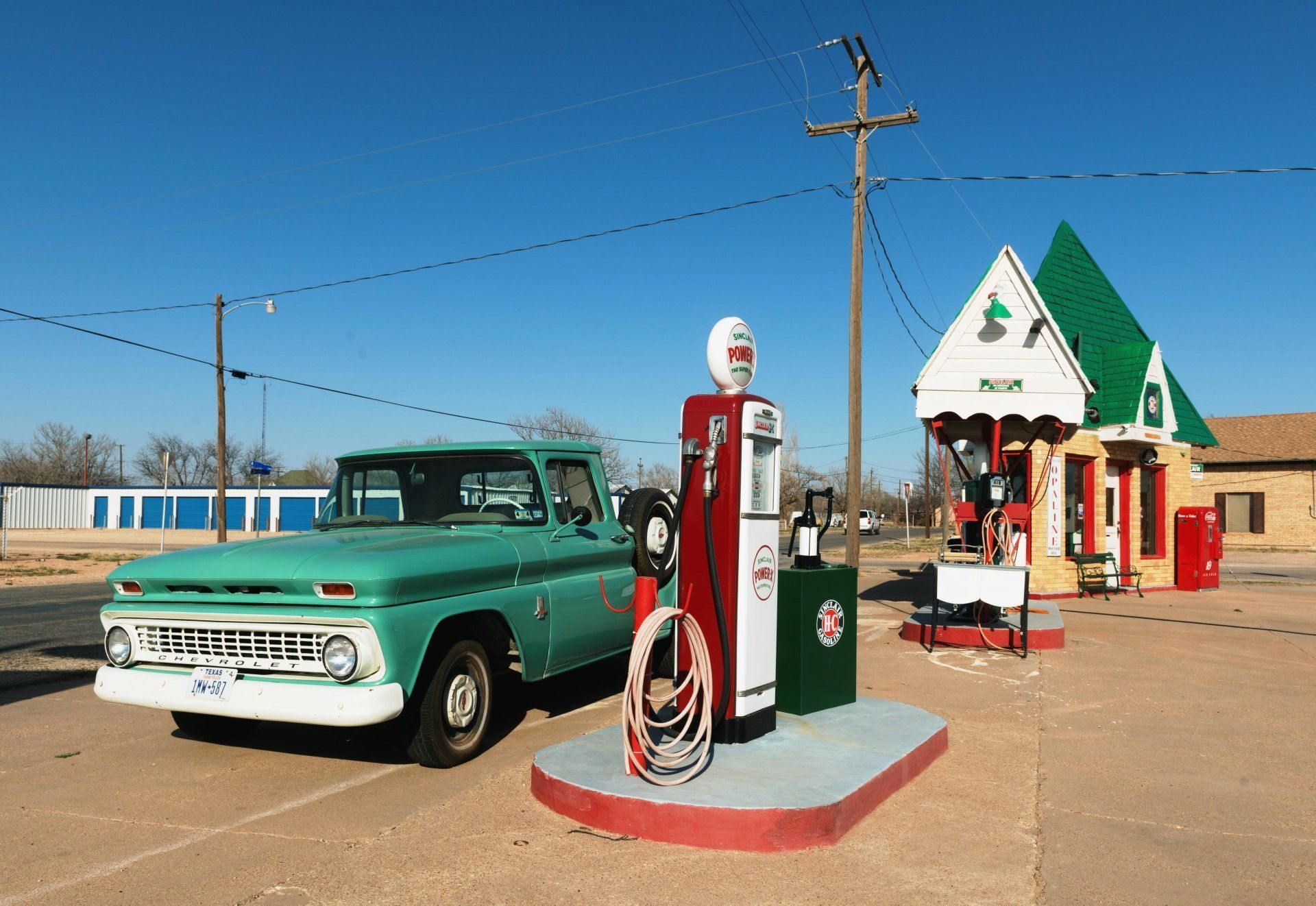 Green vintage pickup truck at an old gas station with red and white pump and green roof.
