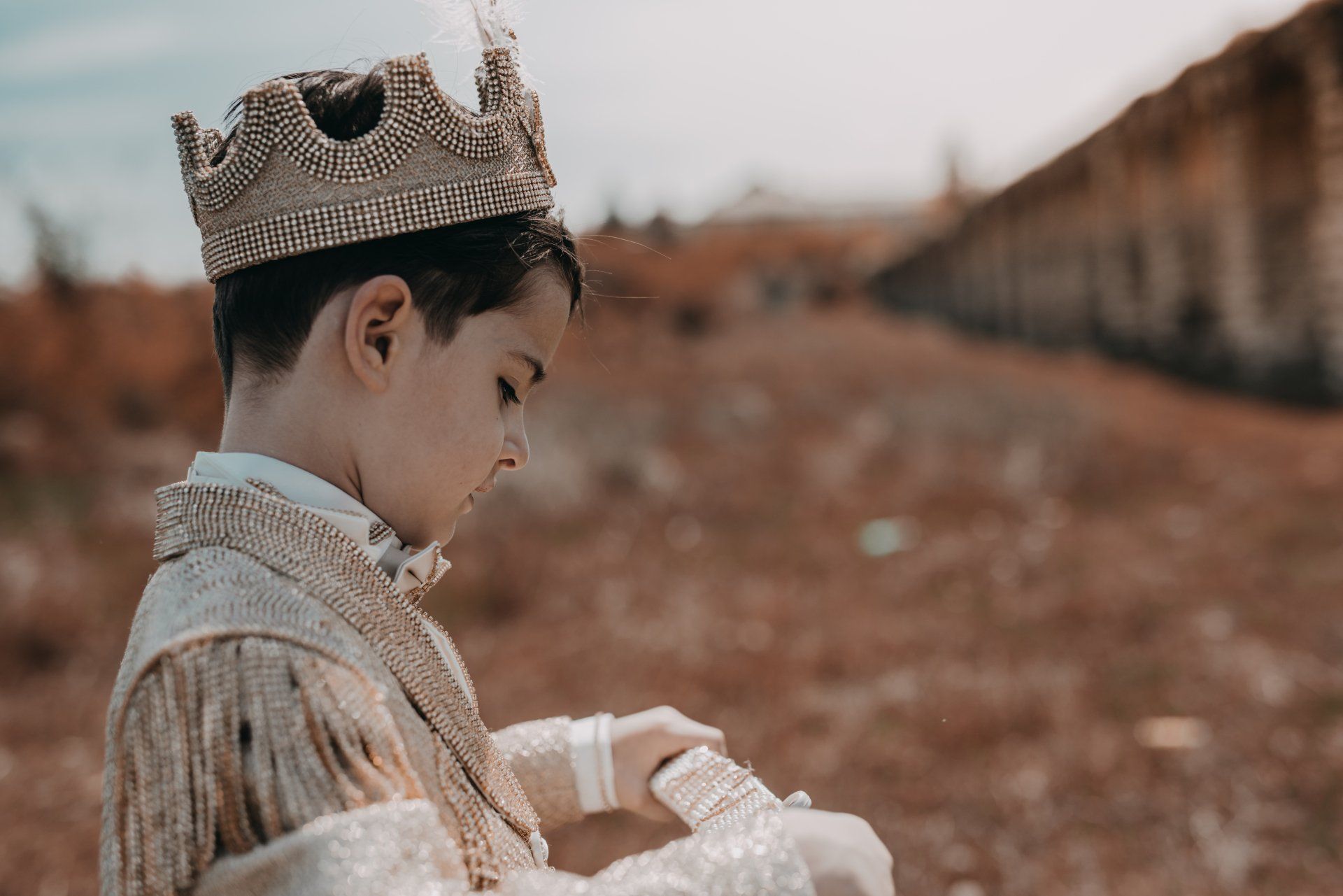 Boy in gold crown and jacket looking down, outdoors.