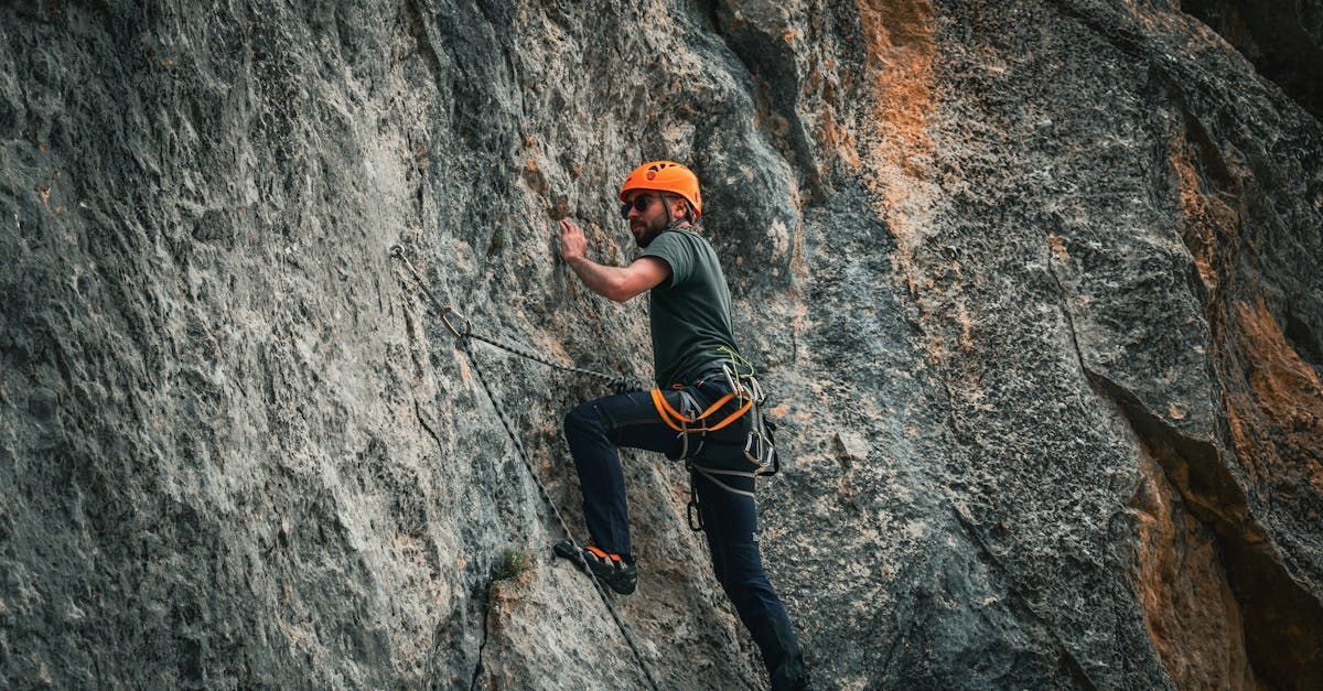 Rock climber in an orange helmet ascending a rock face, wearing a harness and climbing shoes.
