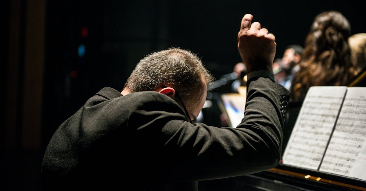 Conductor leading an orchestra with his right arm raised; stage lit, sheet music visible.