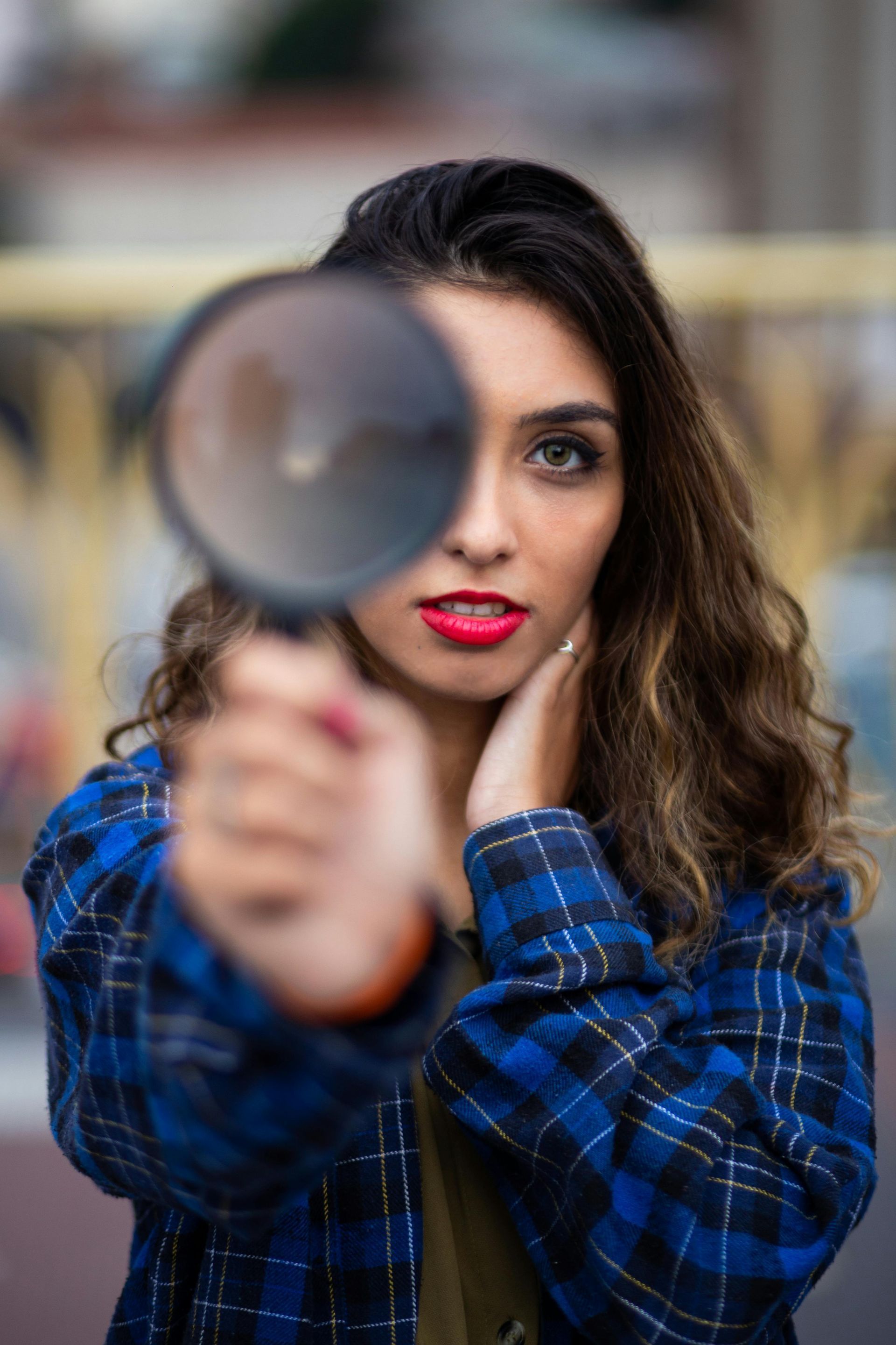 Woman holding magnifying glass, looking intently at camera; wearing blue plaid shirt and red lipstick.