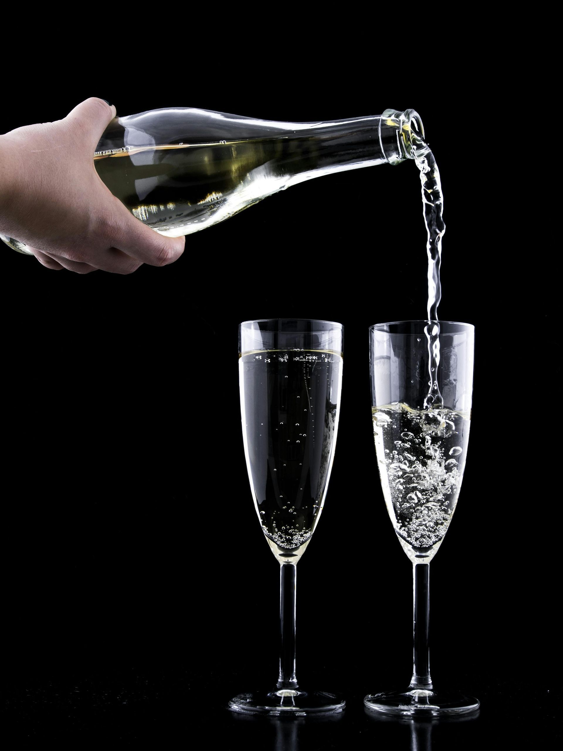 Hand pouring champagne from a bottle into two fluted glasses against a black background.