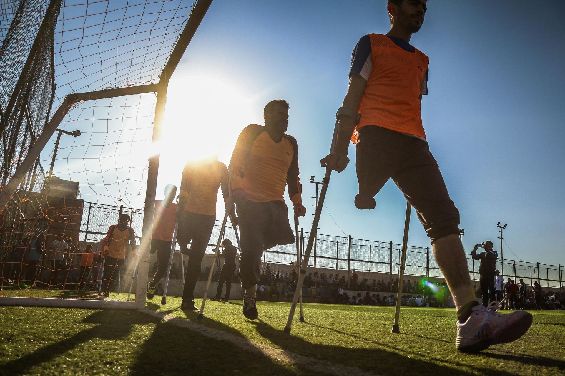 Men on crutches playing soccer on a sunny field; goal in the background.