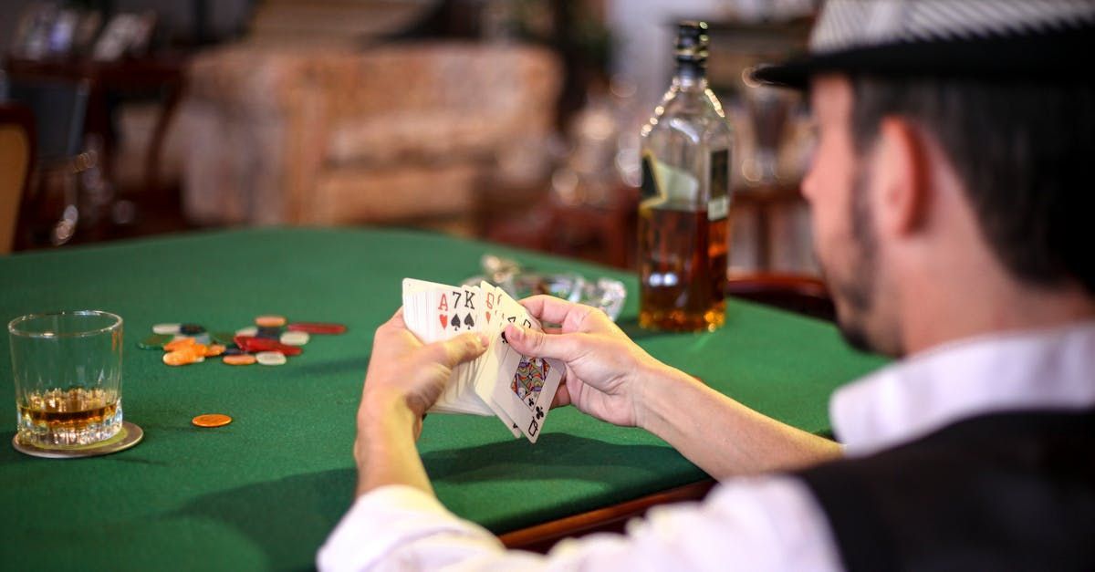 Man in hat holding cards at poker table, glass of whiskey, bottle of liquor, chips on green felt.