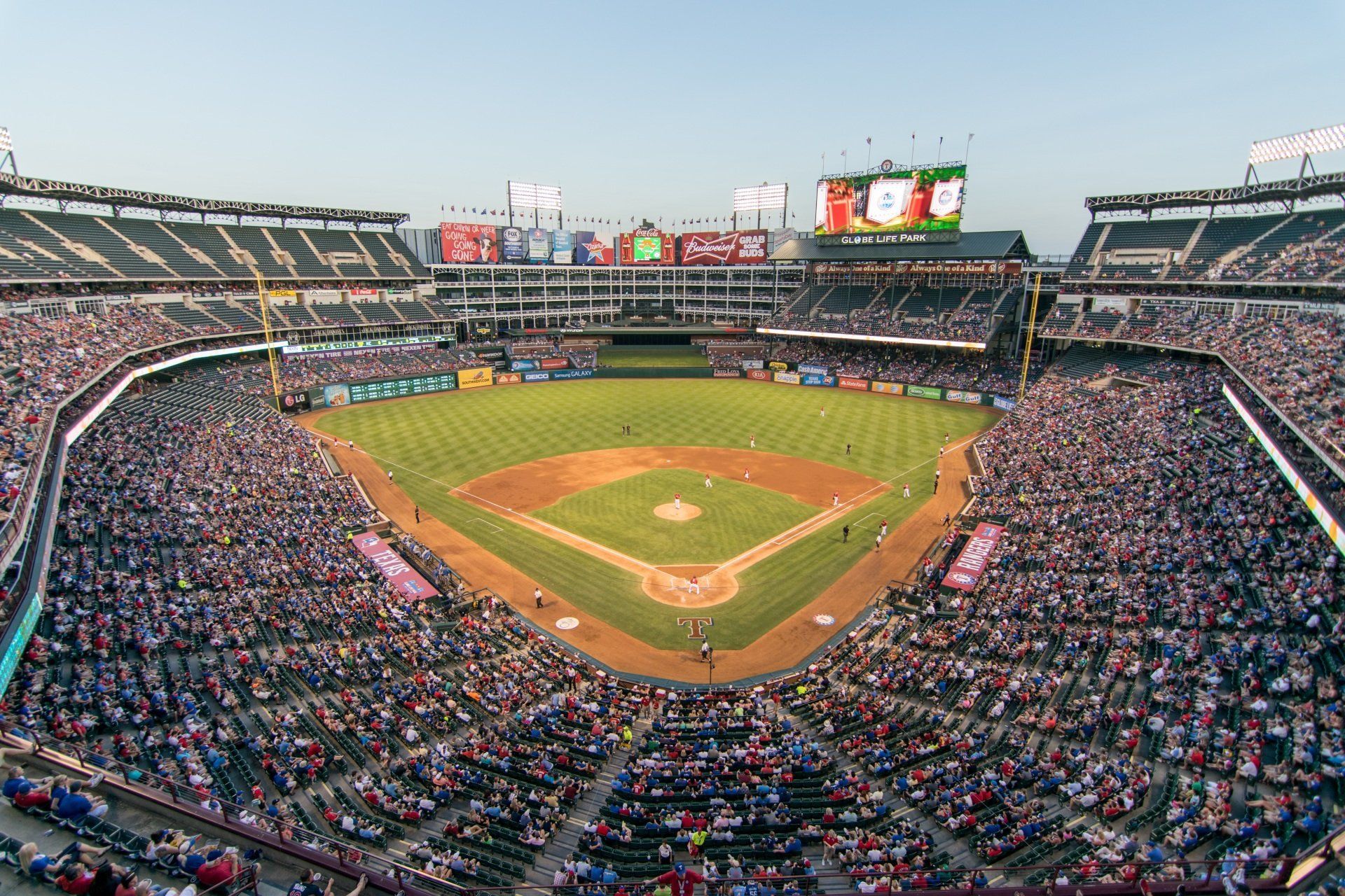 Baseball stadium filled with spectators; green field, brown infield, scoreboard, blue sky.