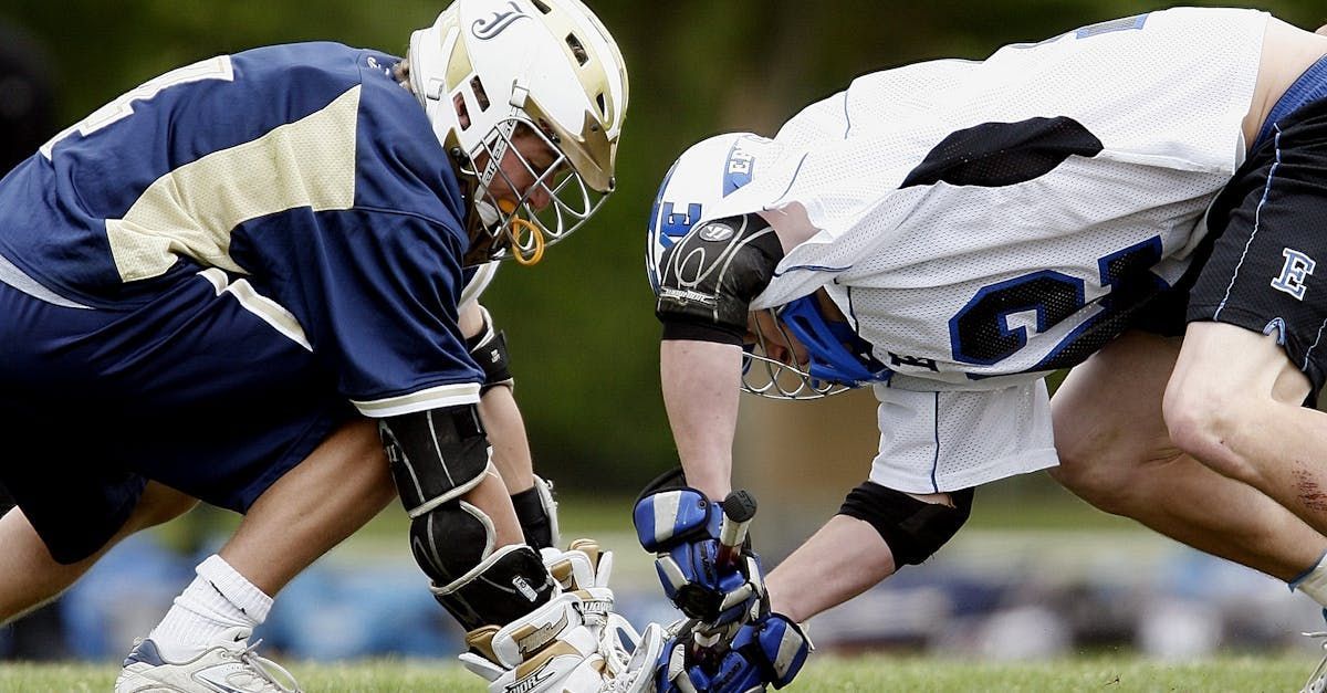 Lacrosse players in dark blue and white uniforms face off on a green field, helmets down.