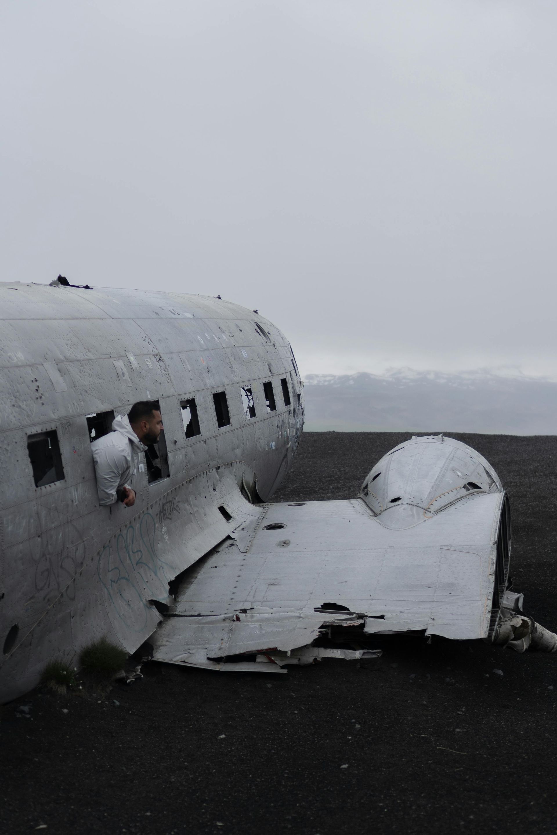 Man looking out of a damaged airplane on black sand beach, overcast sky.
