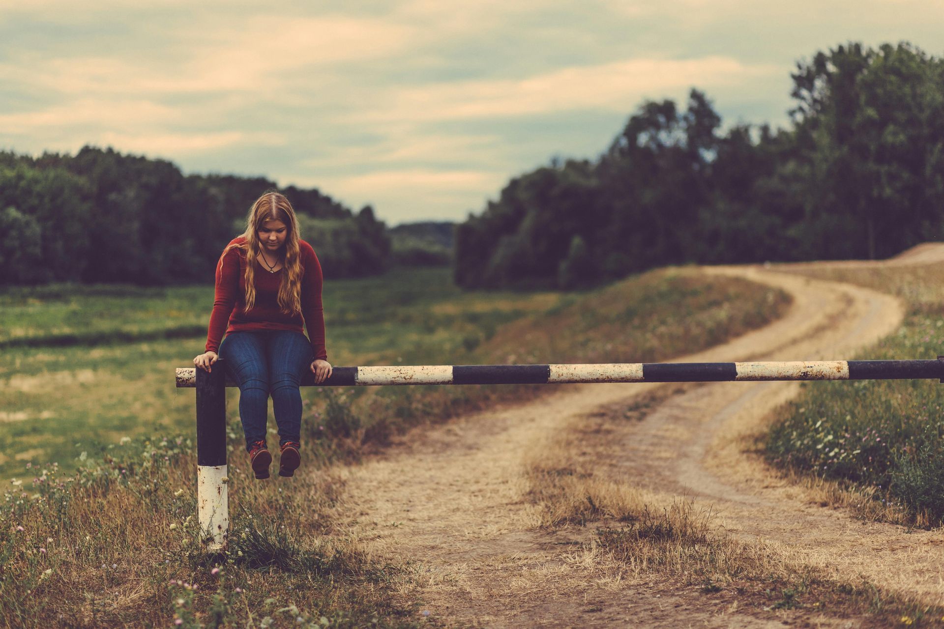 Woman sitting on a barrier at the edge of a dirt road, looking down with a pensive expression in a rural setting.