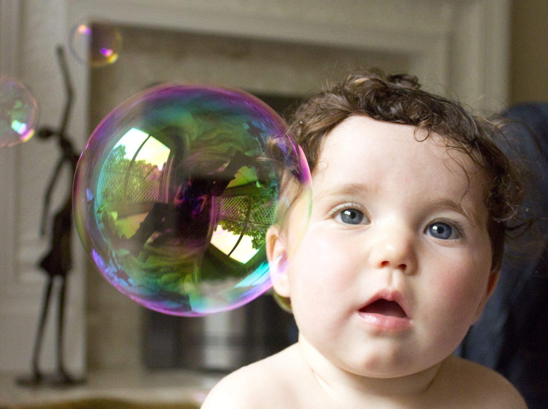 Baby gazes up at a large, iridescent soap bubble near their face, indoors.