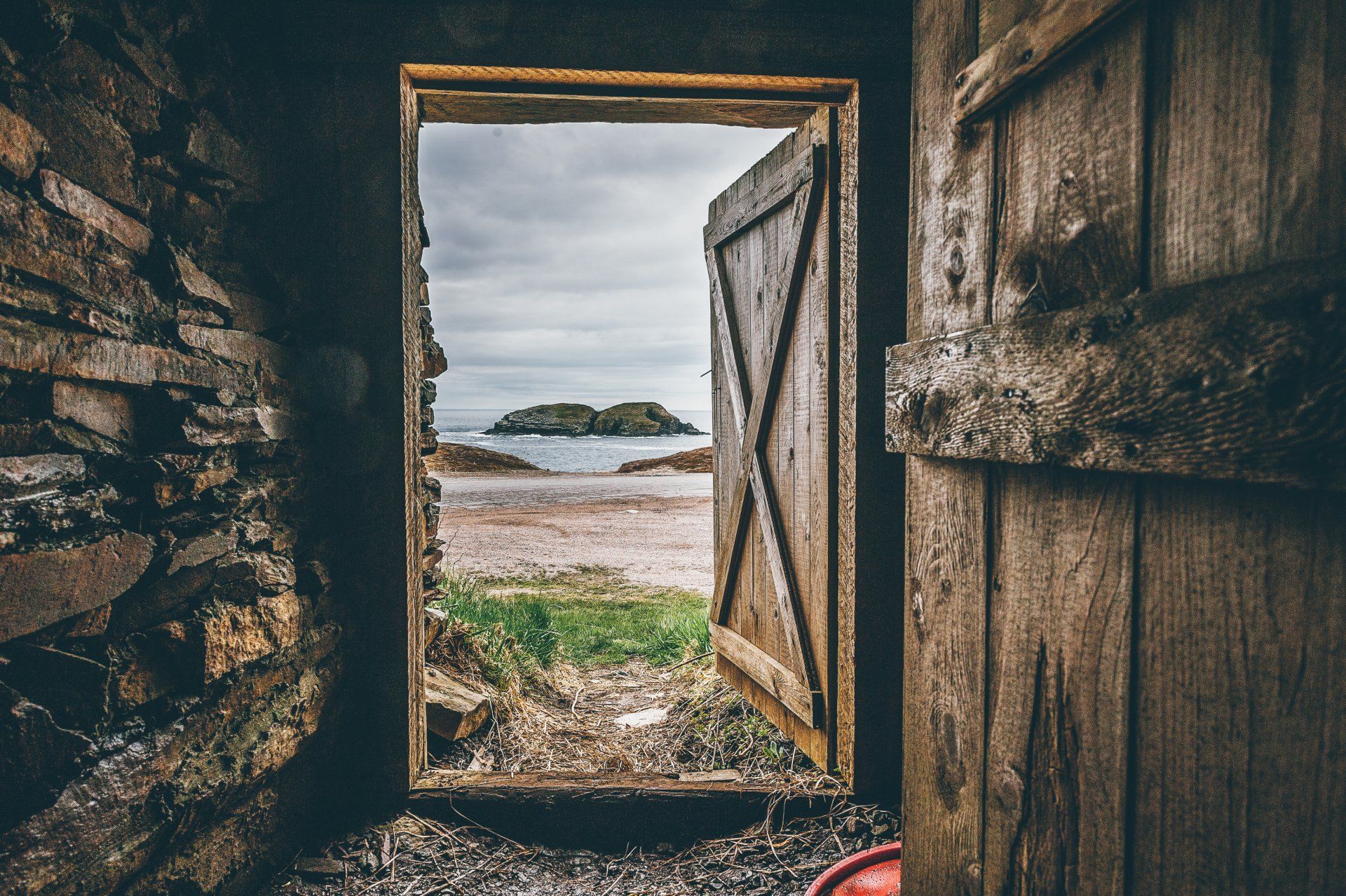 Open wooden door frames a view of a beach, sea, and island under a cloudy sky.