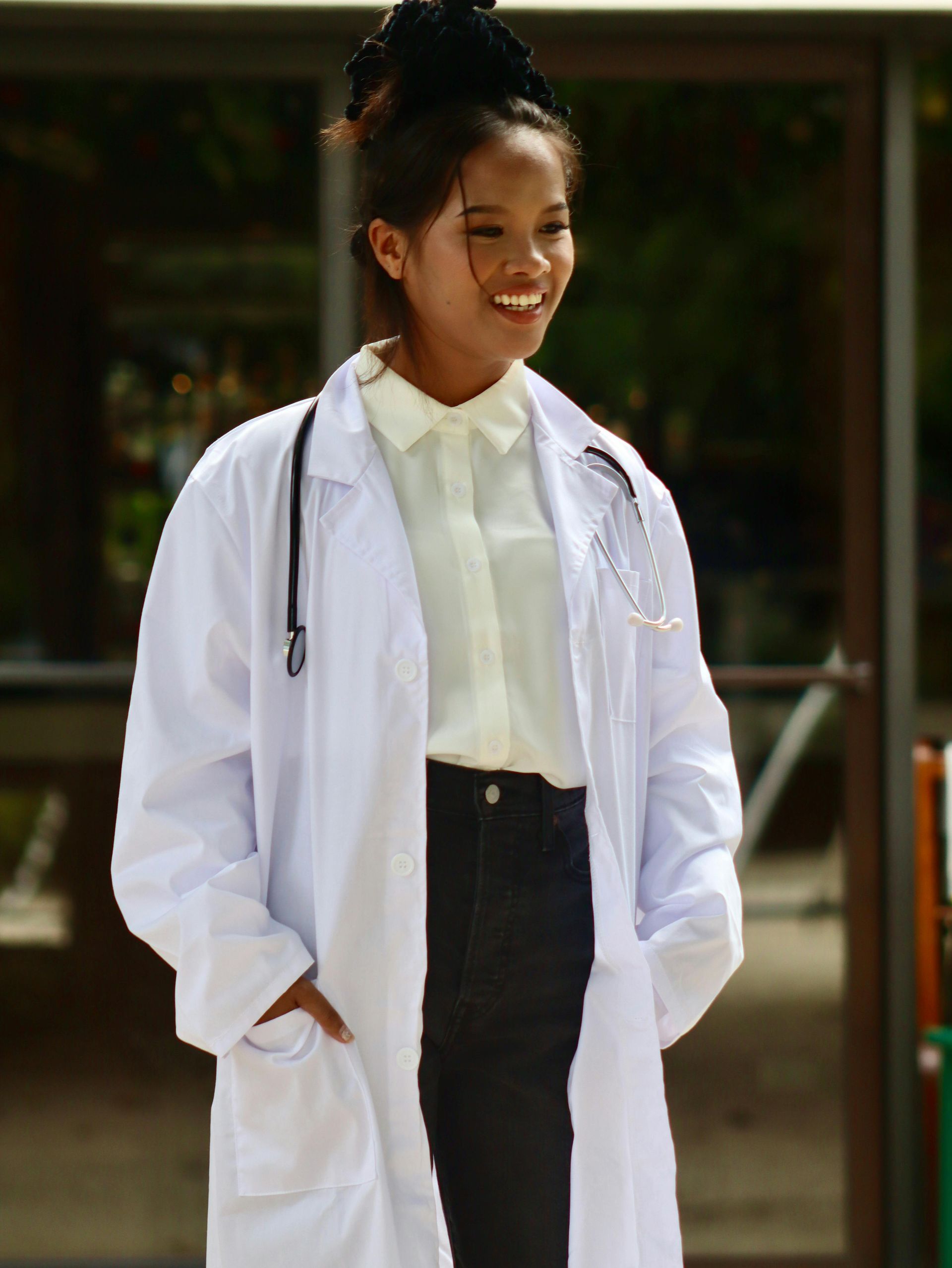 Person in white lab coat with stethoscope, smiles, hands in pockets.