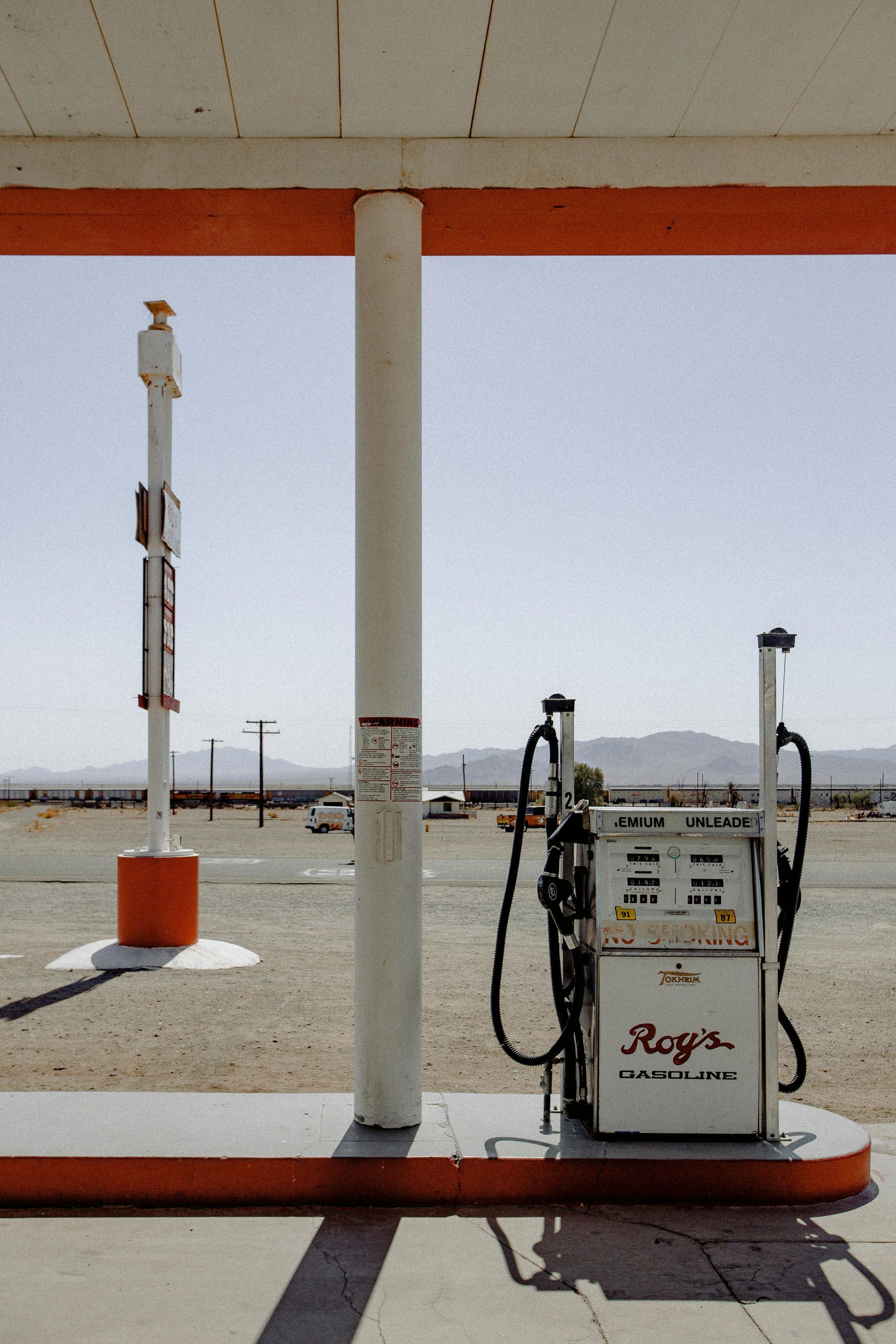 Desert gas station, faded orange and white. Fuel pump, sign, barren landscape, bright sky.
