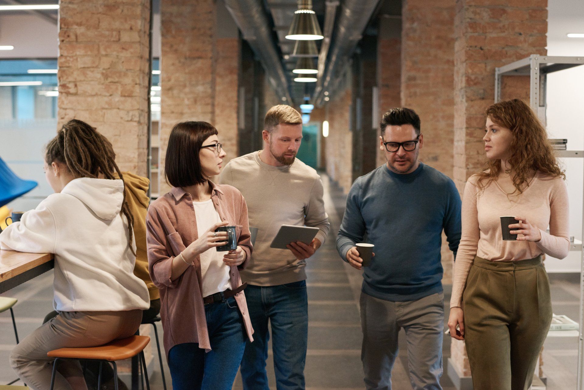 People in office hallway, some holding coffee and a tablet, talking.