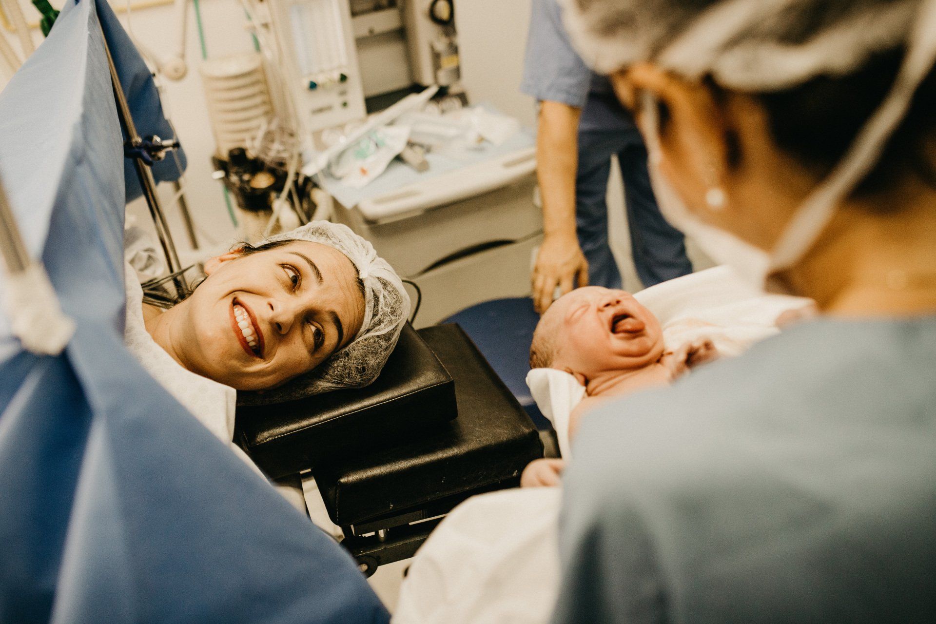 Woman smiles at newborn baby, held by a nurse in a hospital operating room.