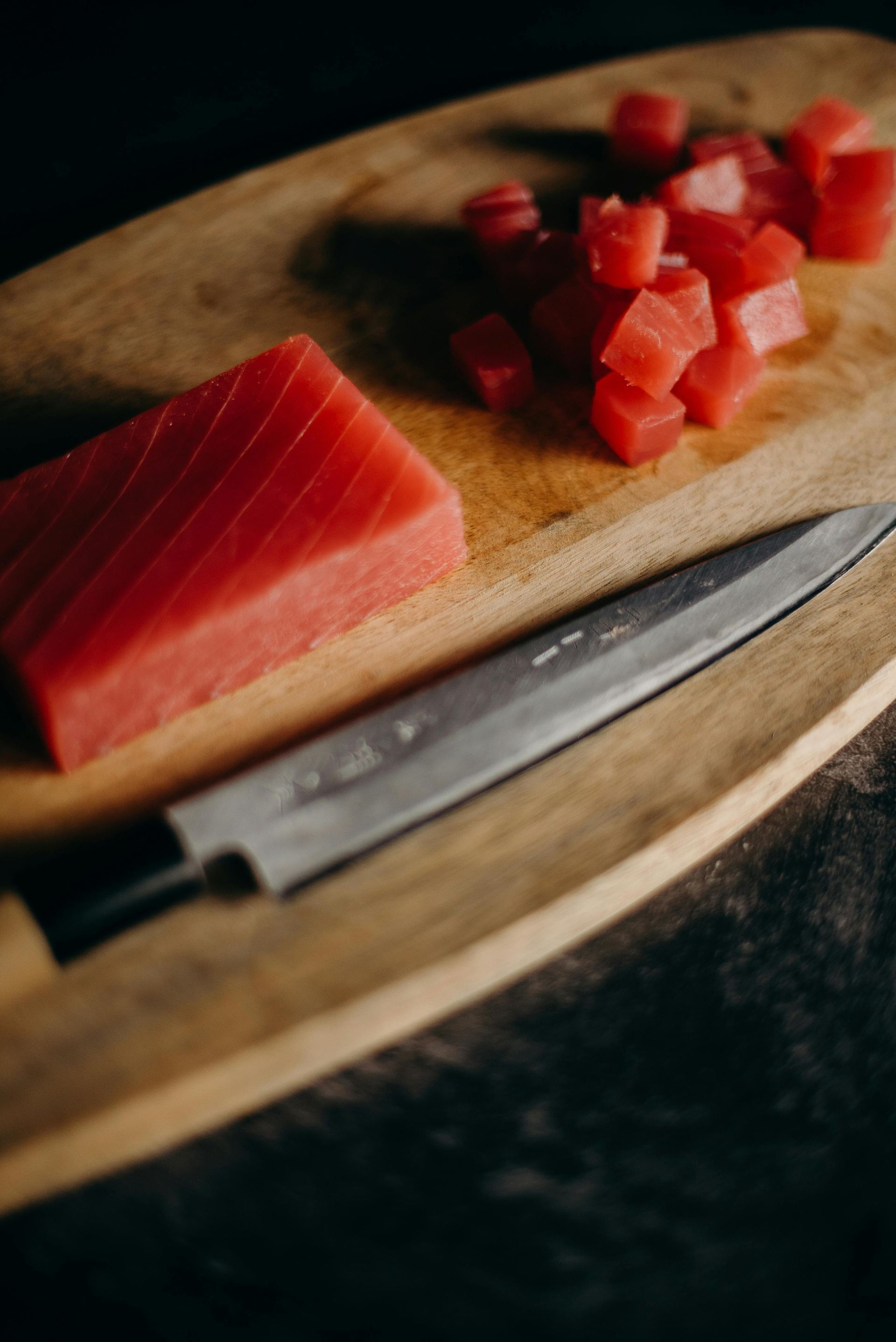 Wooden cutting board with raw tuna, cubed and in a block, next to a knife.