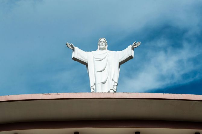 Statue of Jesus with outstretched arms, atop a building, against a cloudy sky.