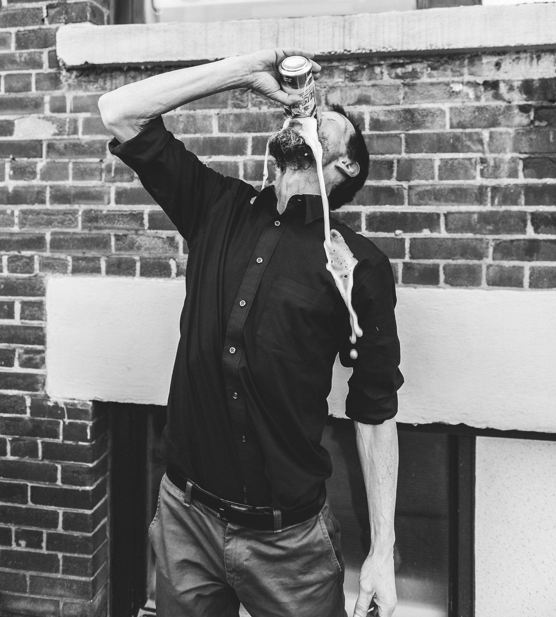 Man drinking, beer overflowing, shirt and face covered in foam, outside by brick wall.