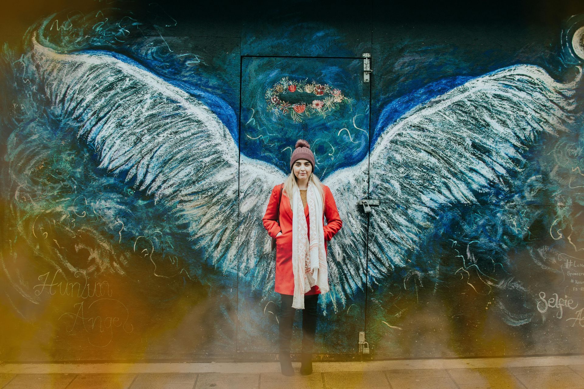 Woman in red coat stands before a mural of angel wings and a door adorned with a wreath.