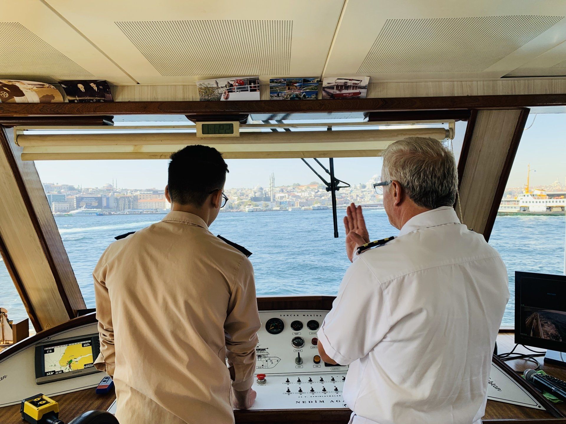Two people in a ship's cockpit looking at the water. One gestures to the view.