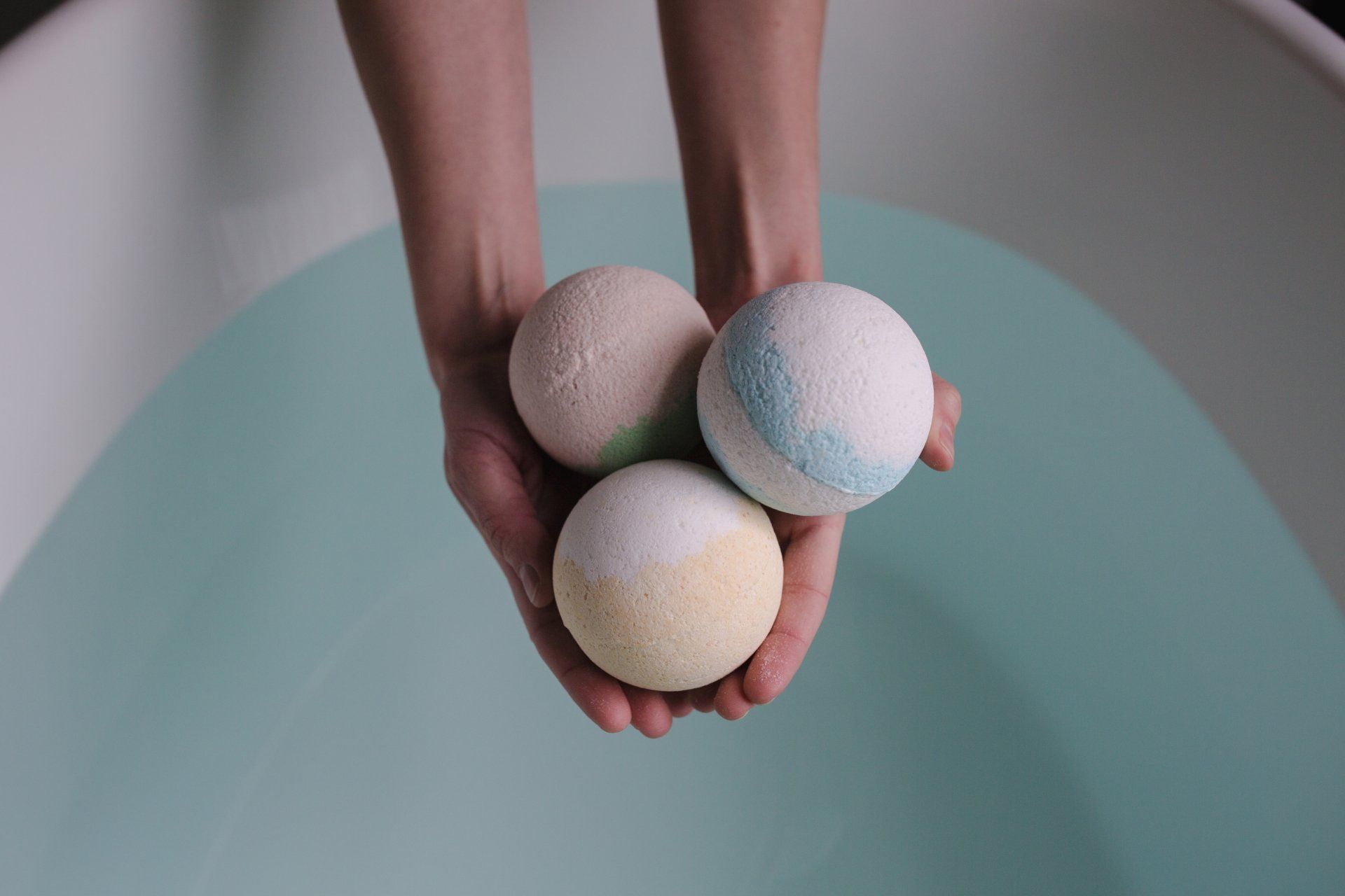 Hands holding three colorful bath bombs over a bathtub filled with light blue water.