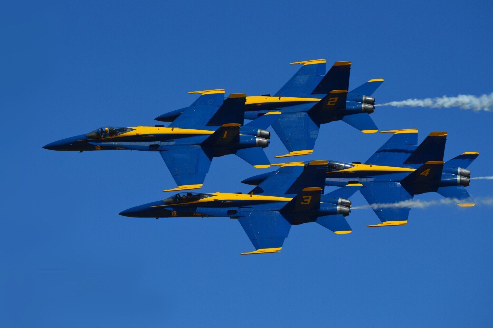 Four blue and yellow jets in formation against a clear blue sky, leaving white contrails.