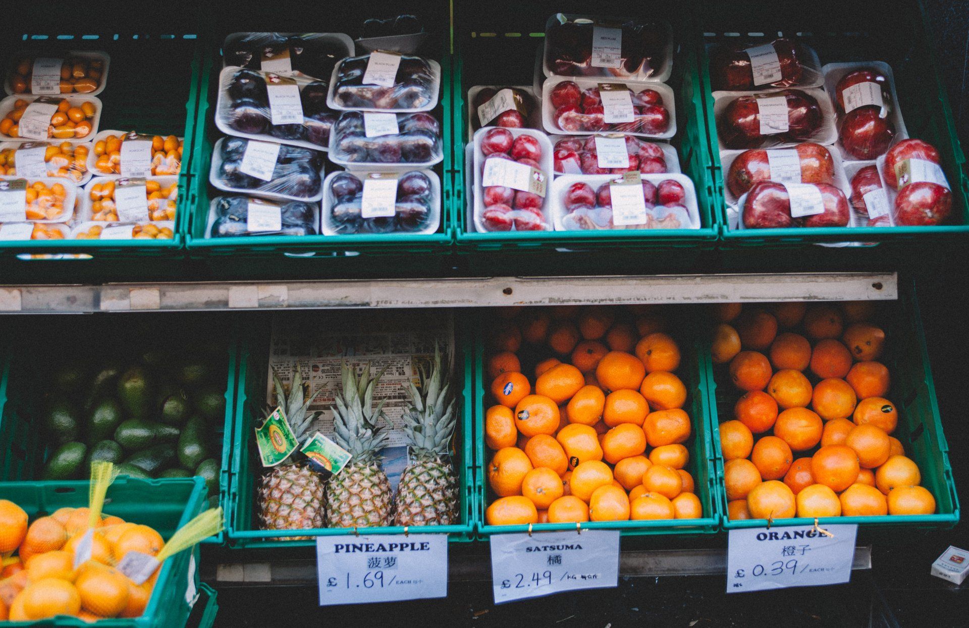 Fruit stand display: Tomatoes, plums, apples, oranges, and pineapples in green crates.
