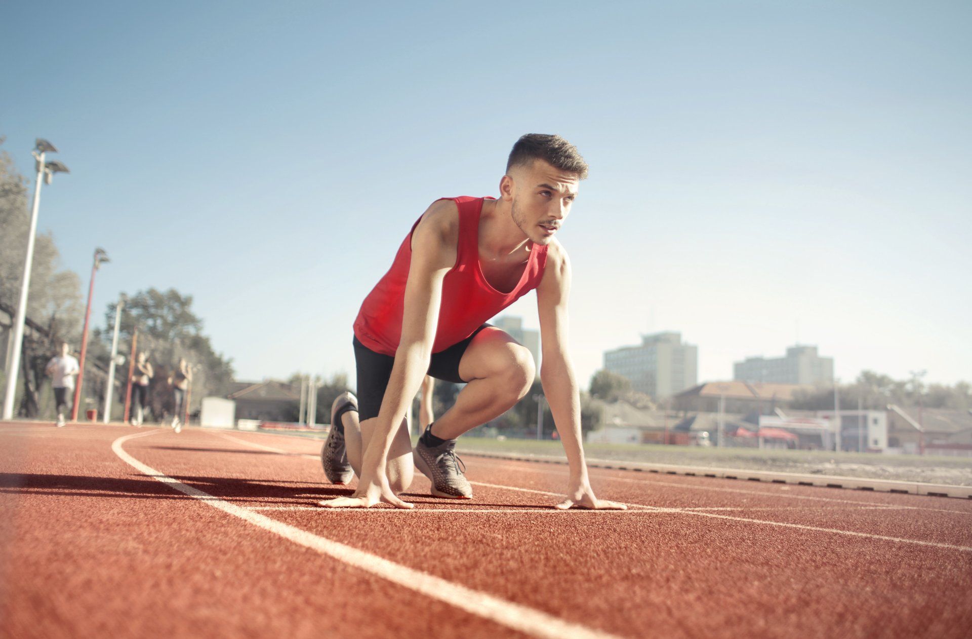 Runner in red tank top in starting position on a track. Blue sky, buildings in the background.
