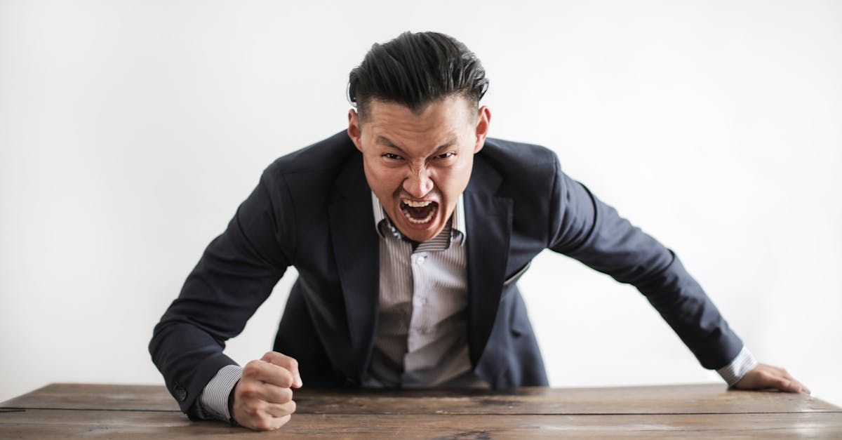Man in suit, angry expression, leaning over a wooden table, fists clenched, white background.