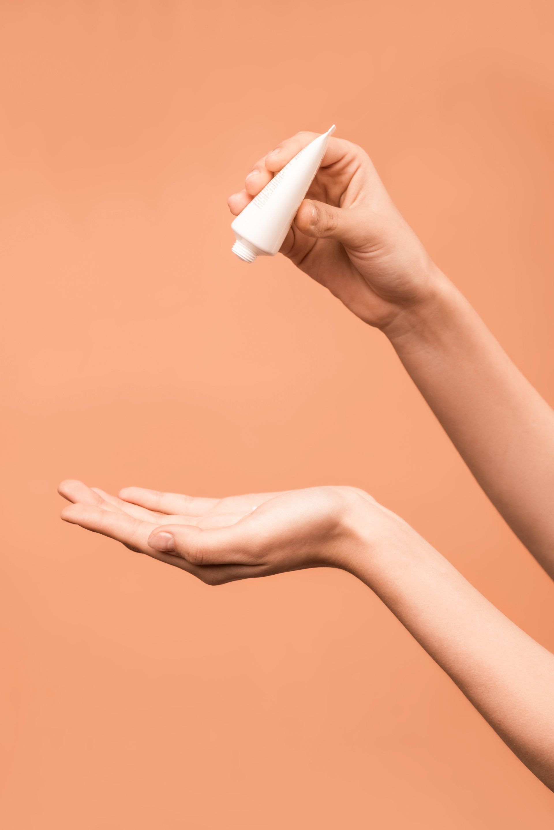 Person squeezing product from a white tube into an open hand, on peach background.