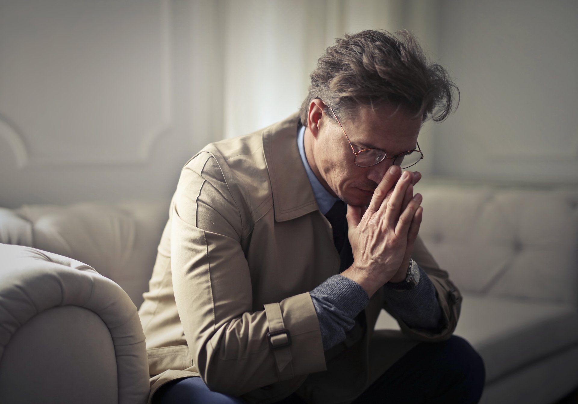 Man in a tan coat, glasses, and suit, sitting with hands clasped, looking down pensively on a white couch.