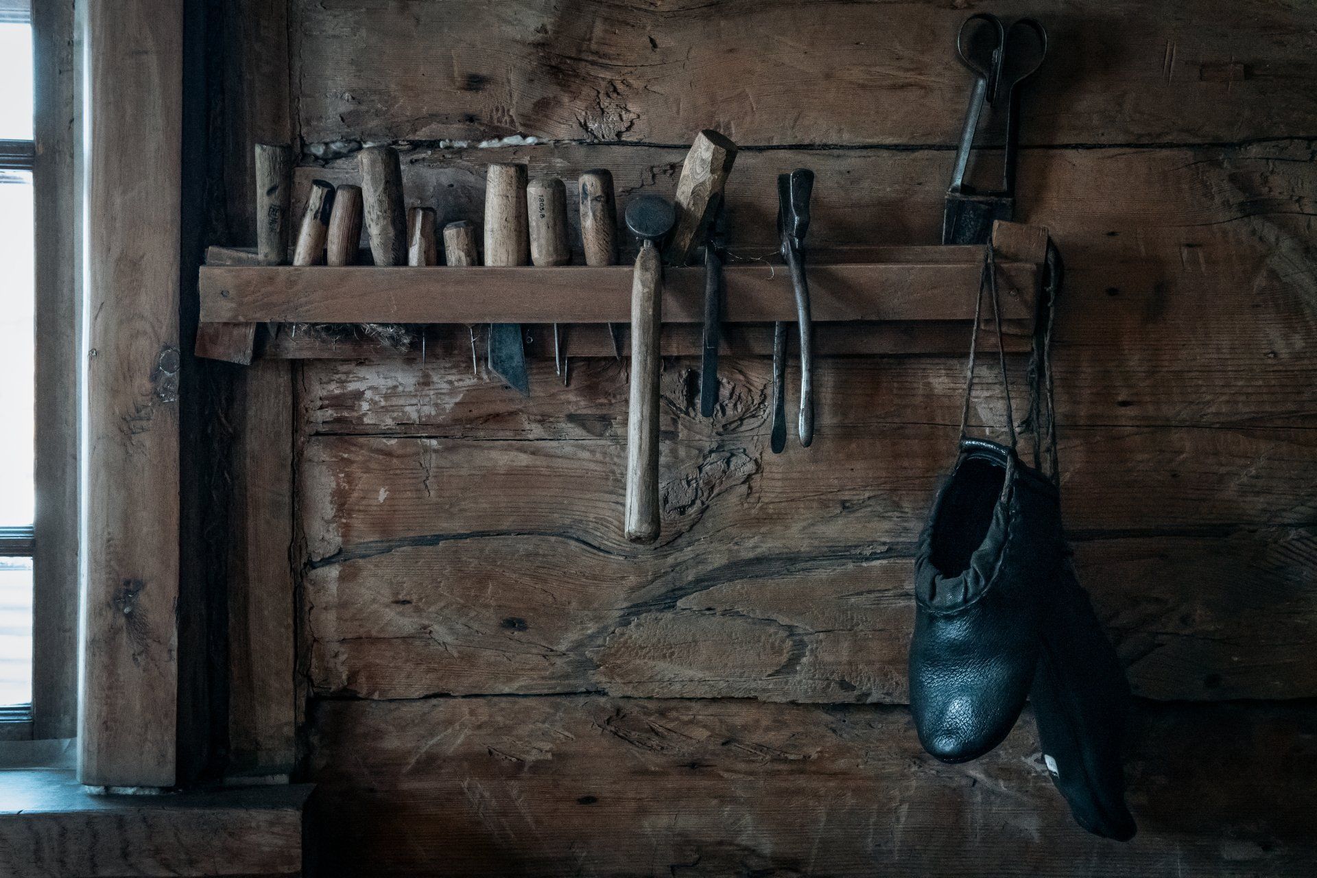 Wooden wall with a shelf holding tools: a hammer, pliers, and various wooden dowels. Two dark shoes hang below.