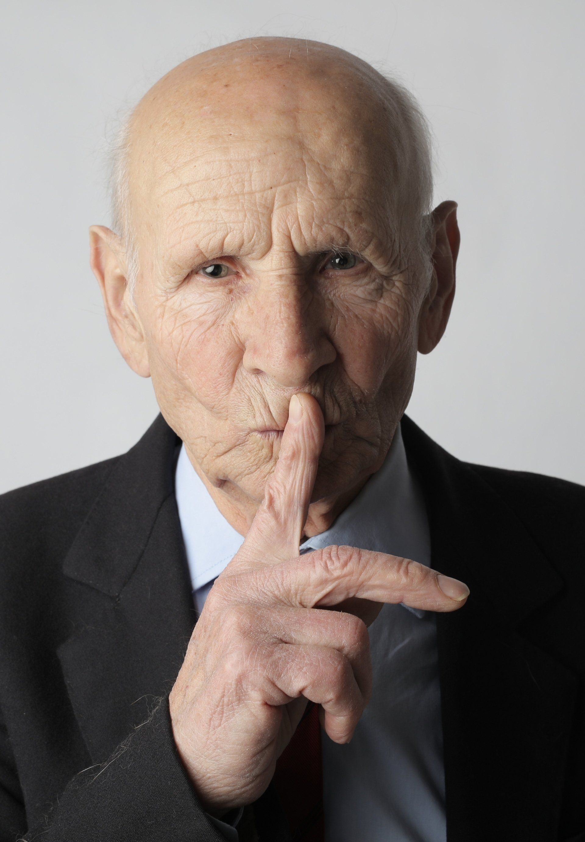 Elderly man in suit, finger to lips, signaling silence.