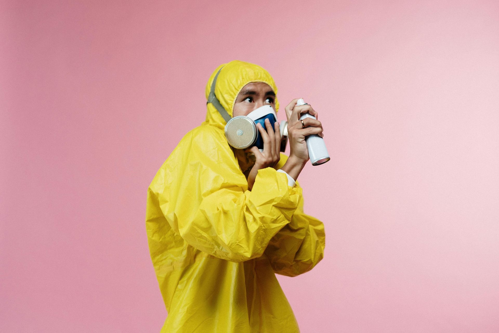 Person in yellow hazmat suit, respirator, and holding spray can, looking off-camera against a pink backdrop.