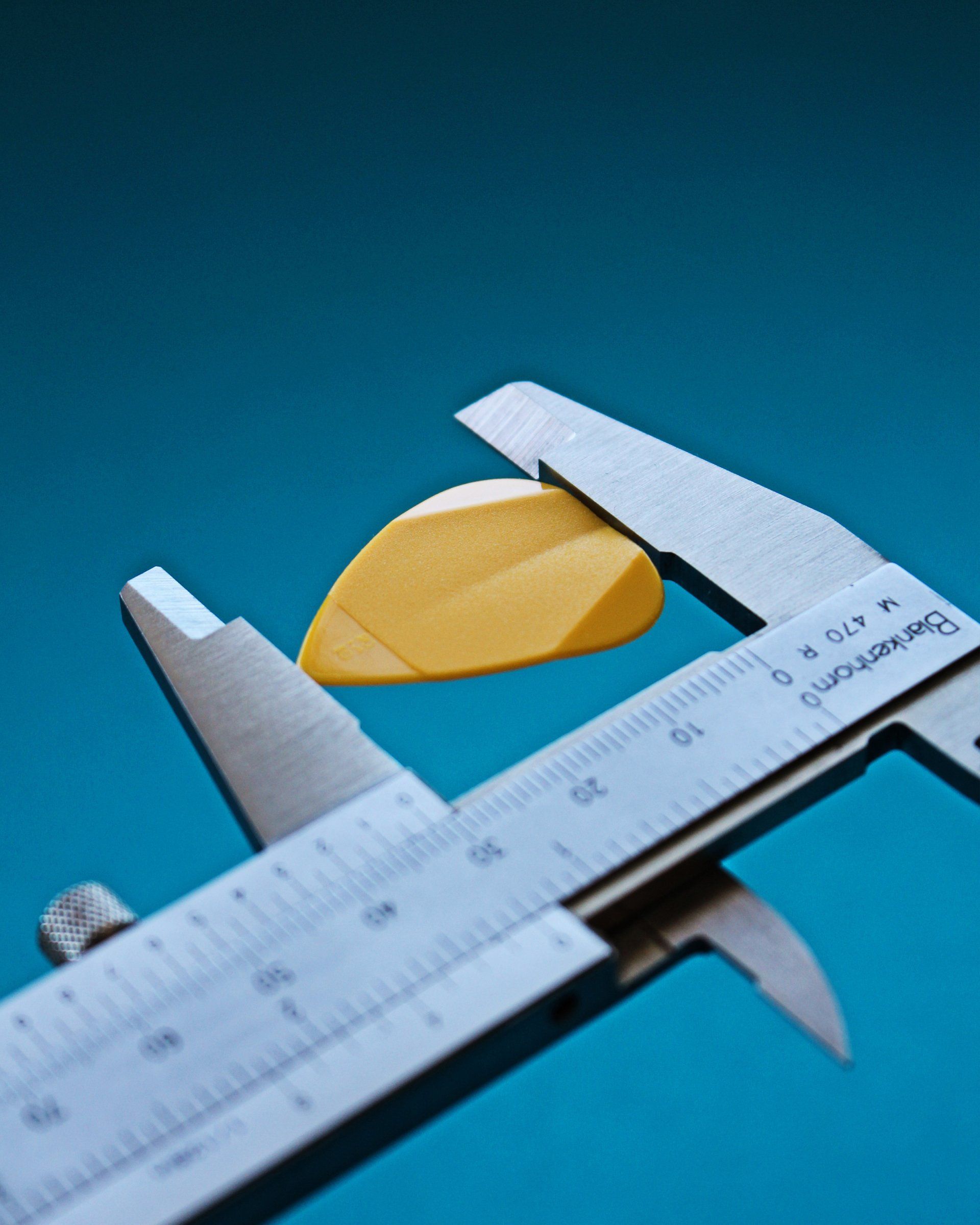 A yellow guitar pick is measured with a silver caliper against a blue background.