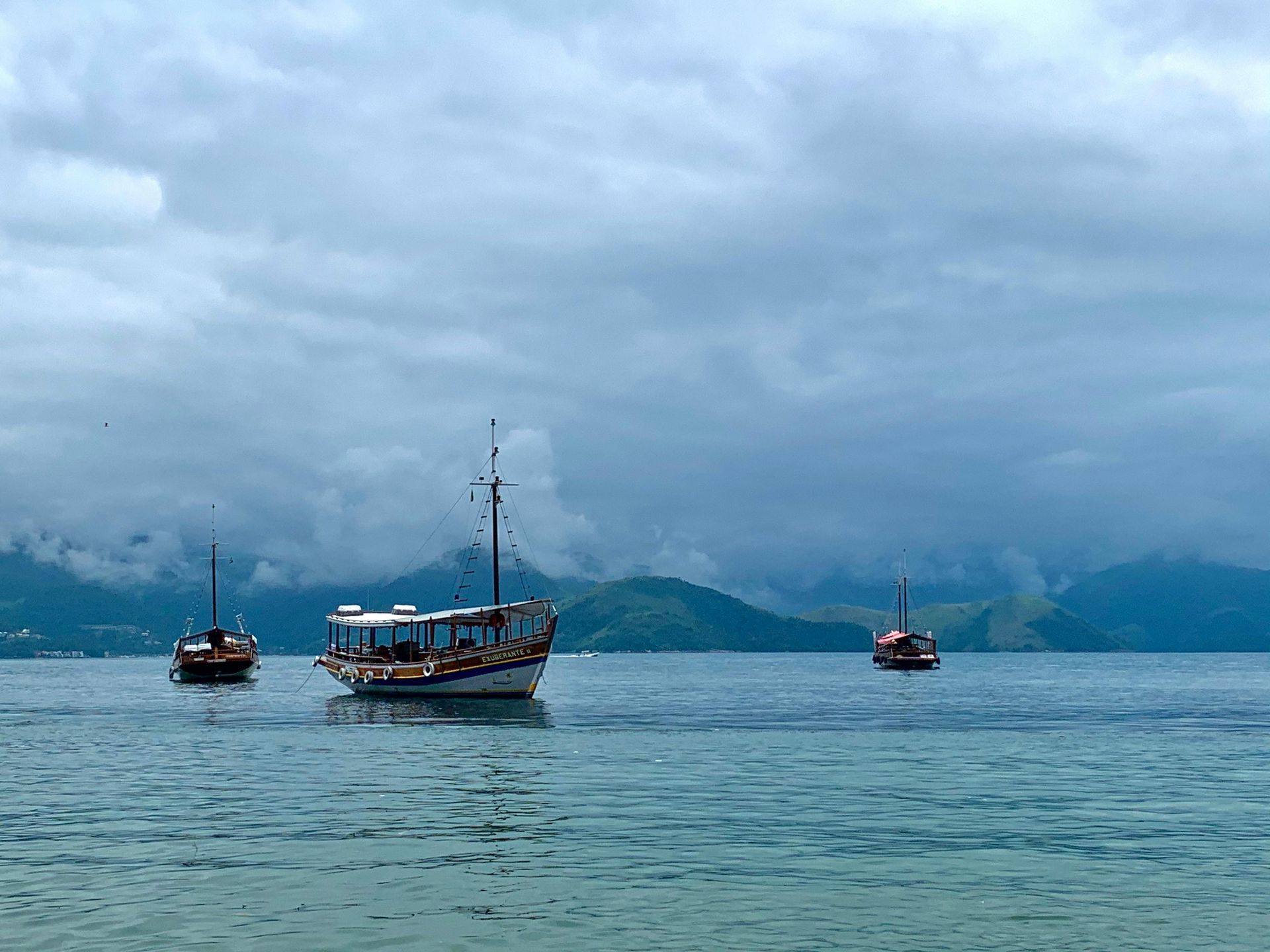 Three boats on calm water under a cloudy sky near a mountainous shoreline.