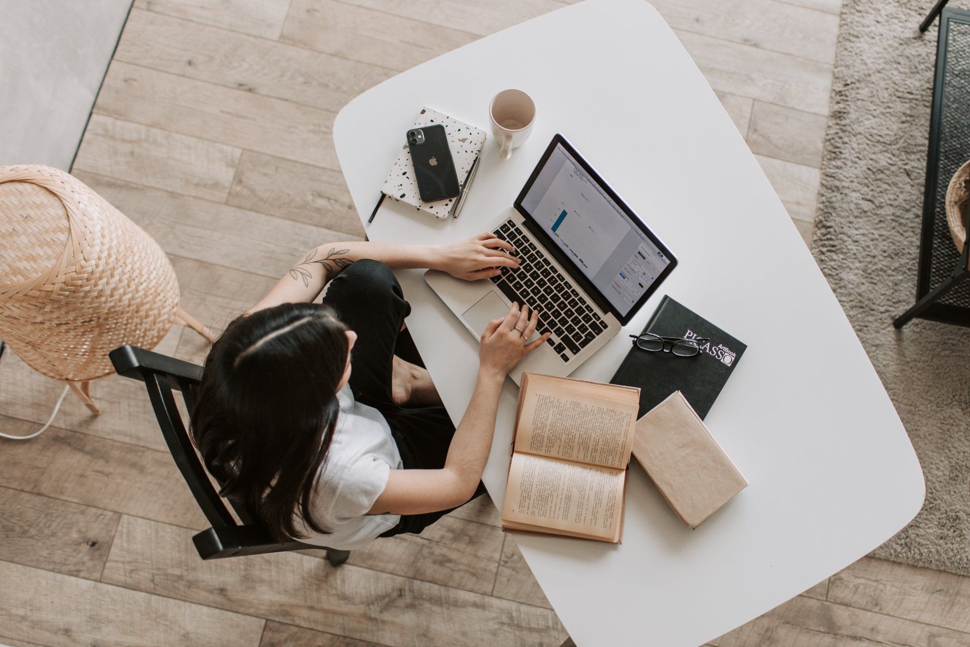 Woman working on laptop at white desk with books and coffee.