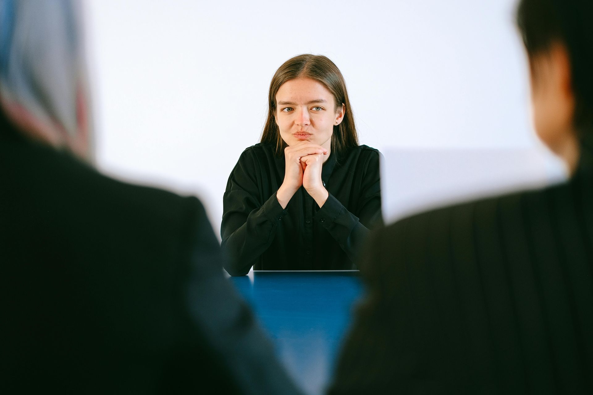 Woman in black shirt, hands clasped, at a table facing two people with backs to the camera.