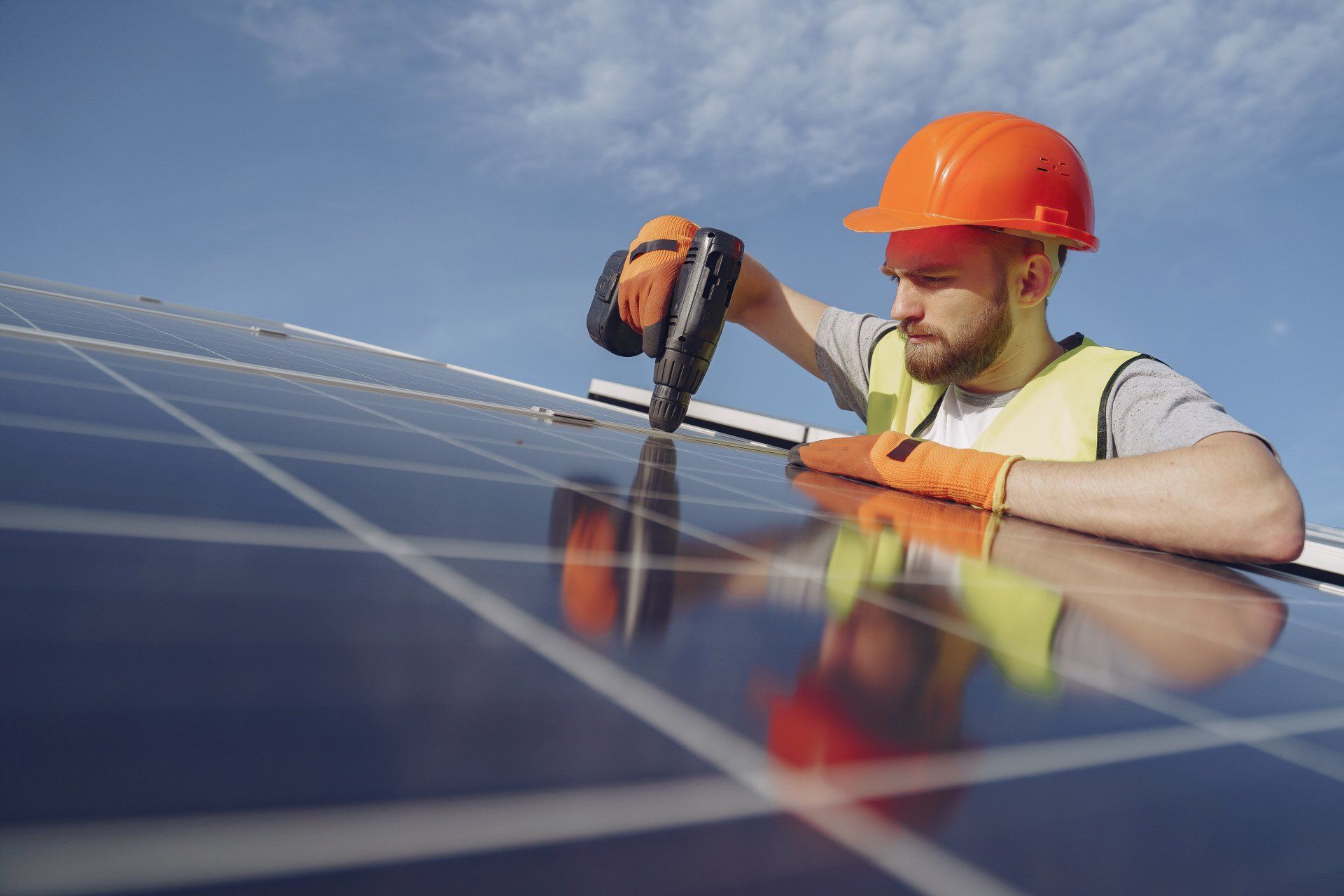 Solar panel installer using a drill on a panel, wearing safety gear against a blue sky.