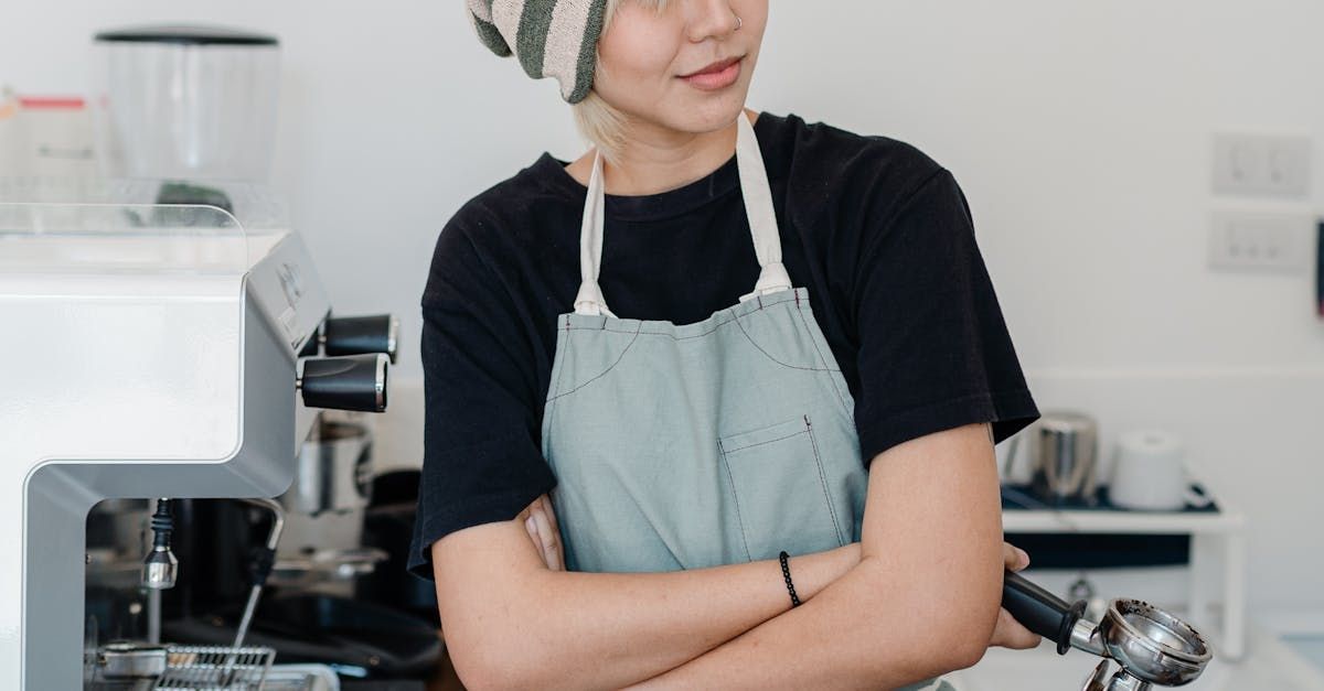 Barista in gray apron and striped beanie with arms crossed, near espresso machine.