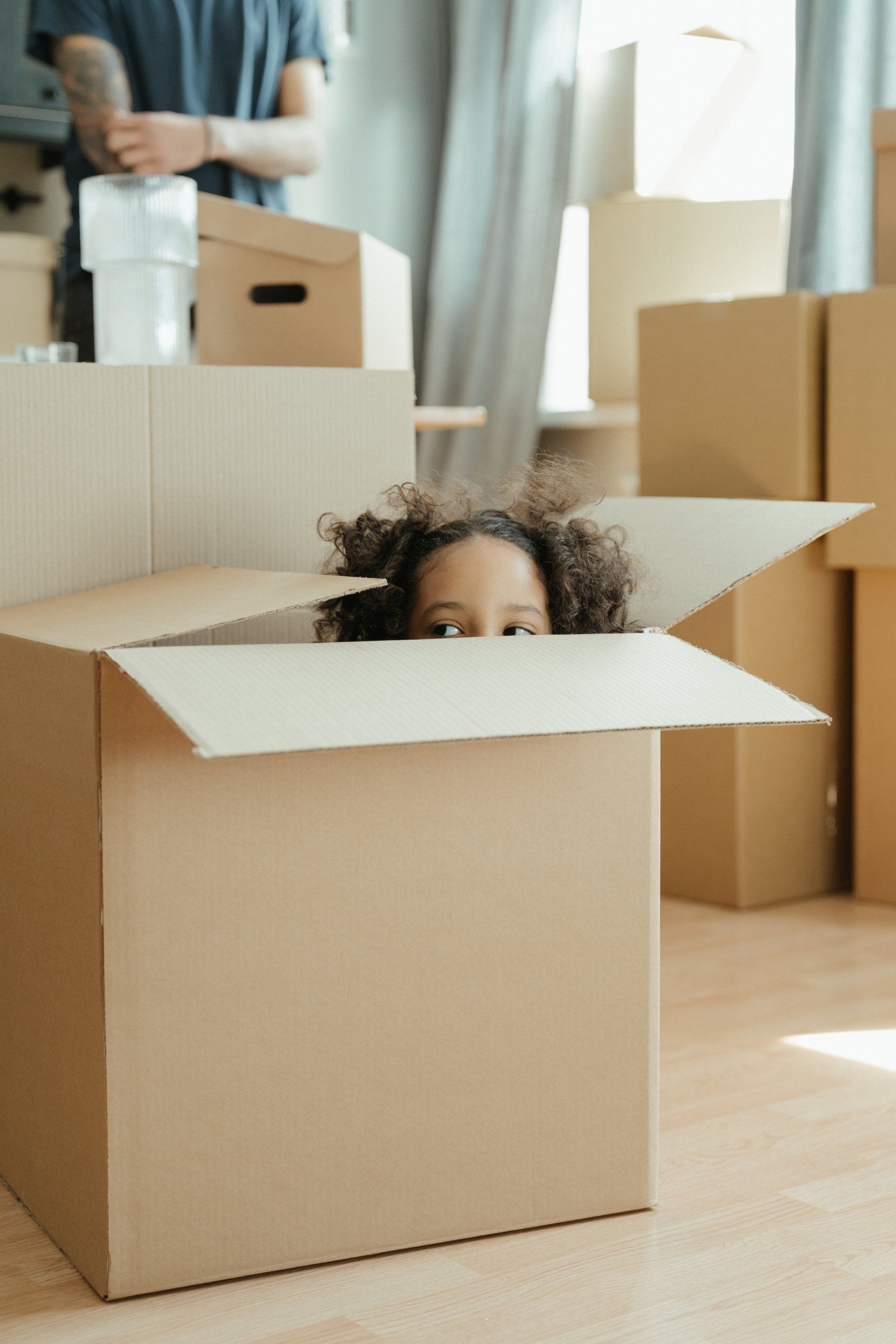 Child peeks out of a cardboard box in a room with other boxes; person in background.