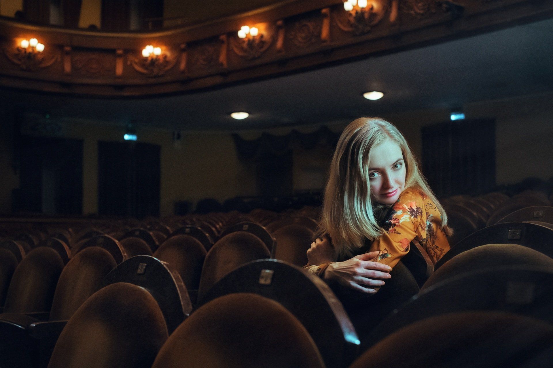 Woman in patterned shirt, smiling in an empty theater auditorium with rows of seats and ornate ceiling lights.