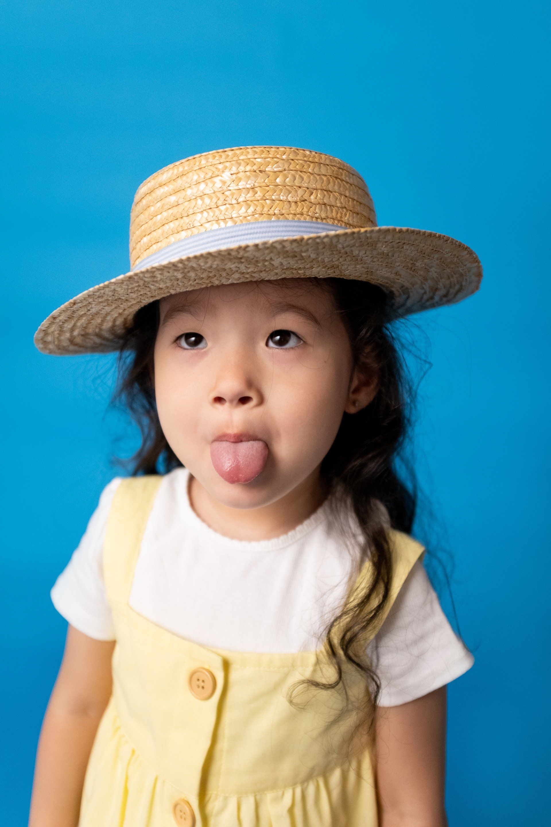 Child sticking tongue out, wearing a straw hat and yellow dress, against a blue background.