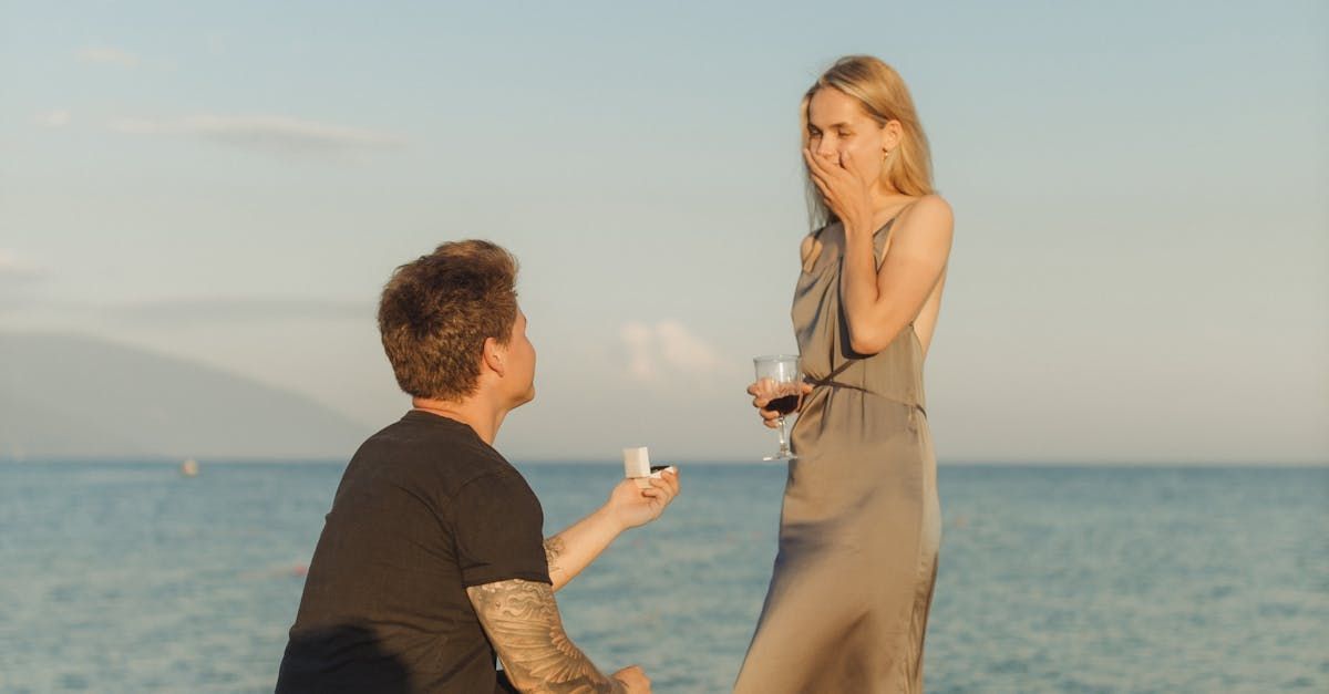 Man kneeling, proposing to woman on beach; she holds wine glass, surprised.