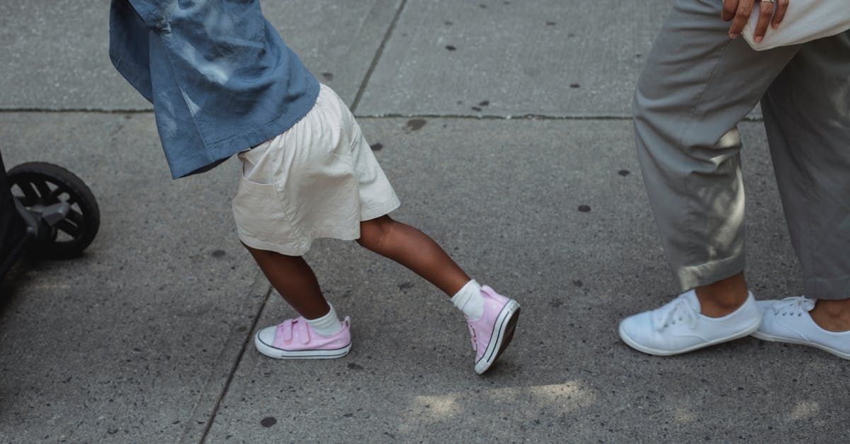 Child in pink shoes walks on sidewalk with adult wearing white shoes.